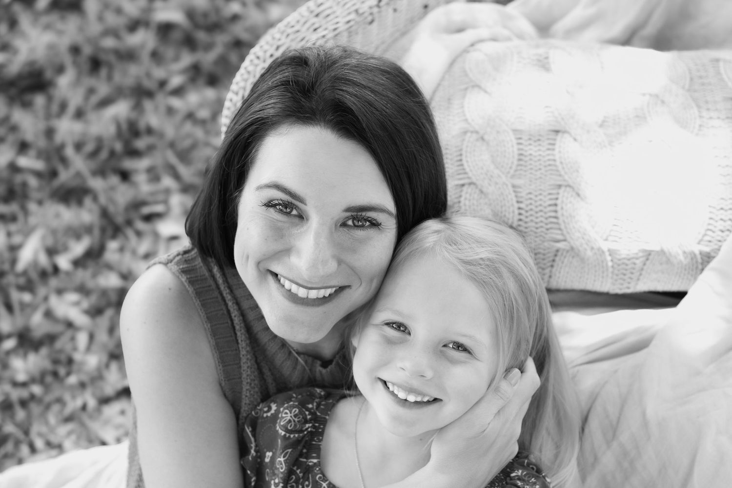 Black and white photo of a smiling mother and young blonde daughter cuddling together outdoors.