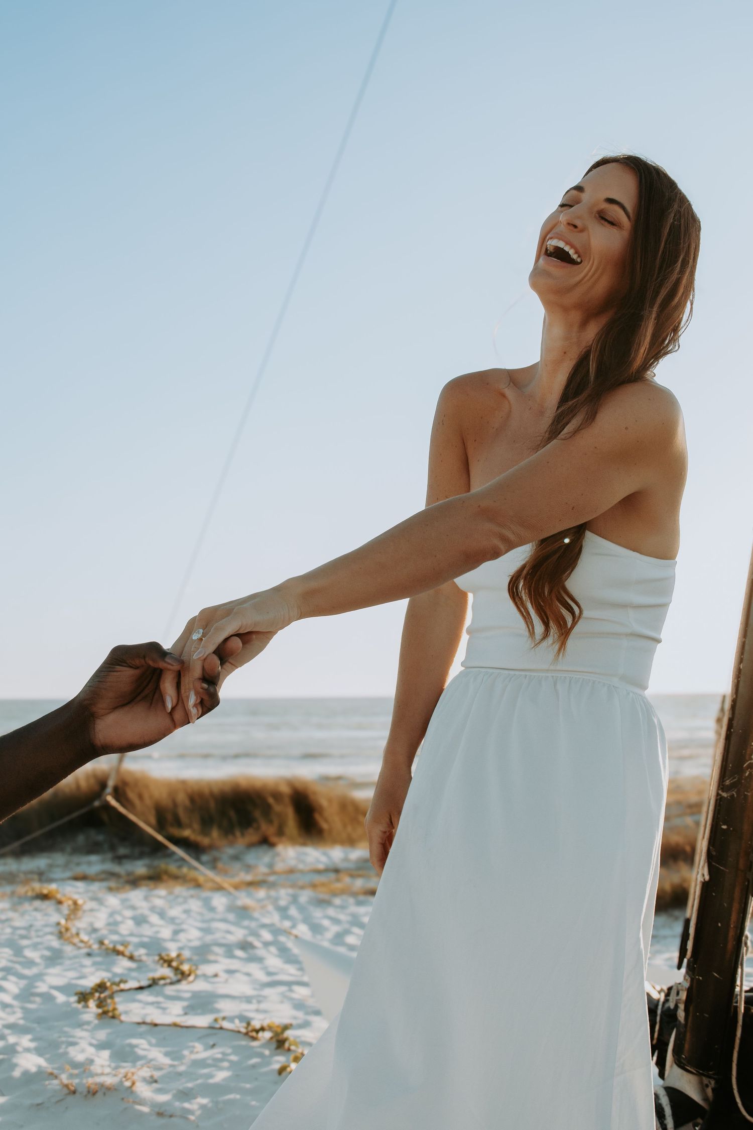 A joyful bride in a white strapless dress laughs while holding hands on a sunny beach.