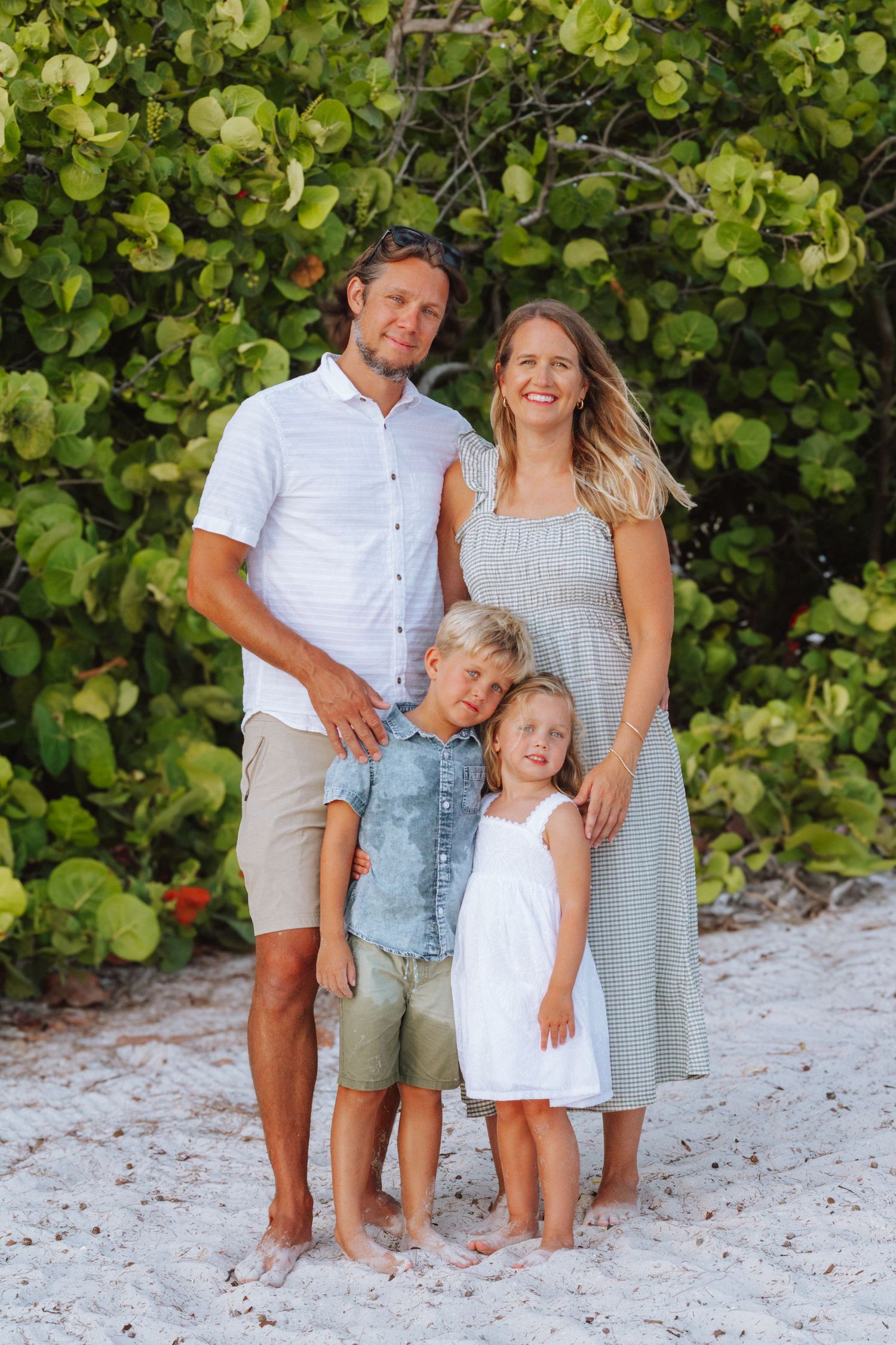 Smiling family of four posing together on white sand beach with tropical green foliage.