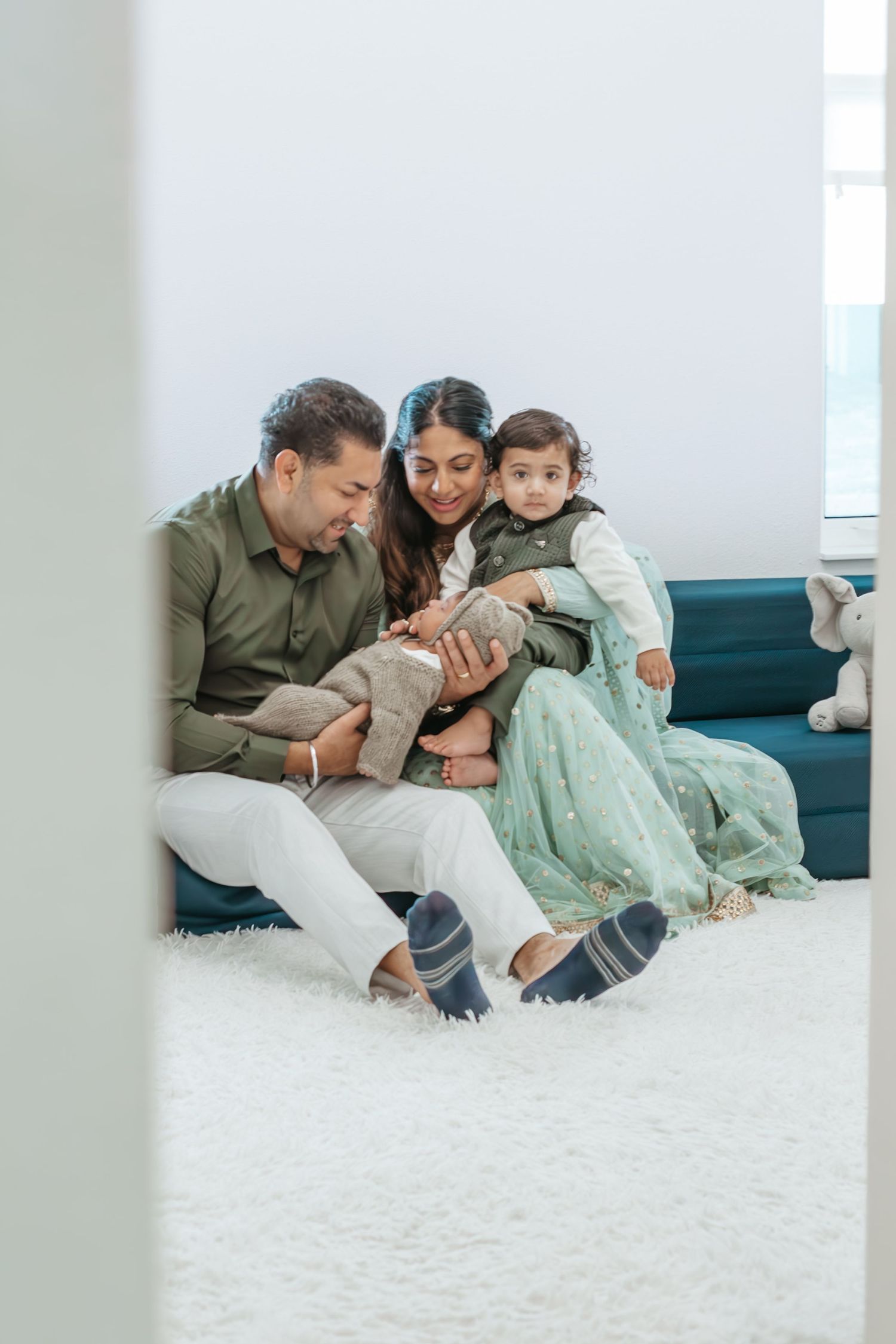 A happy family of three sits together on a teal sofa, parents smiling at their toddler in a bright white room.