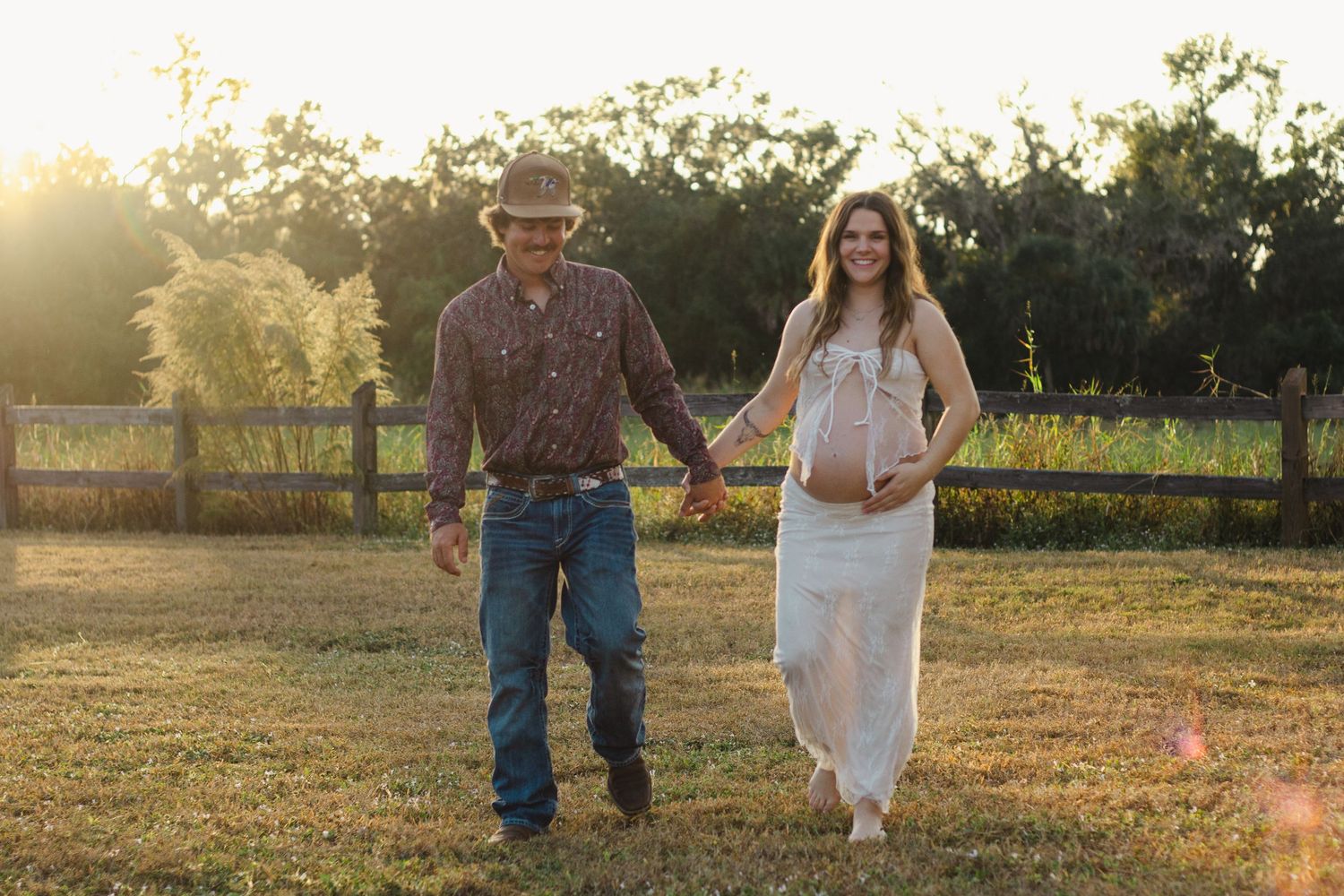 Pregnant couple holding hands in a sunlit field, man in cowboy hat and jeans, woman in white lace maternity dress.