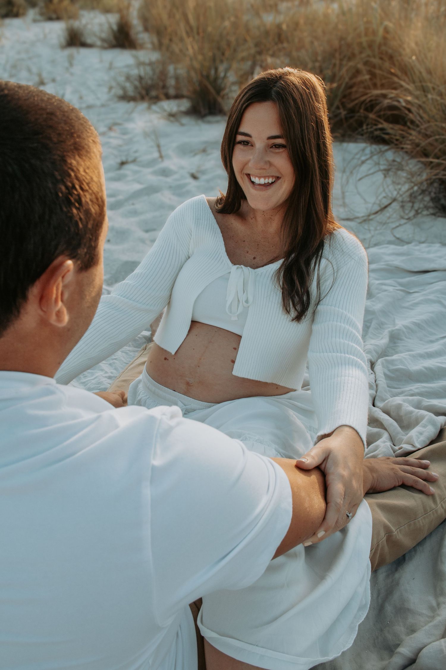 A smiling woman in a white crop top and skirt sits on a beach blanket holding hands with a man in white.