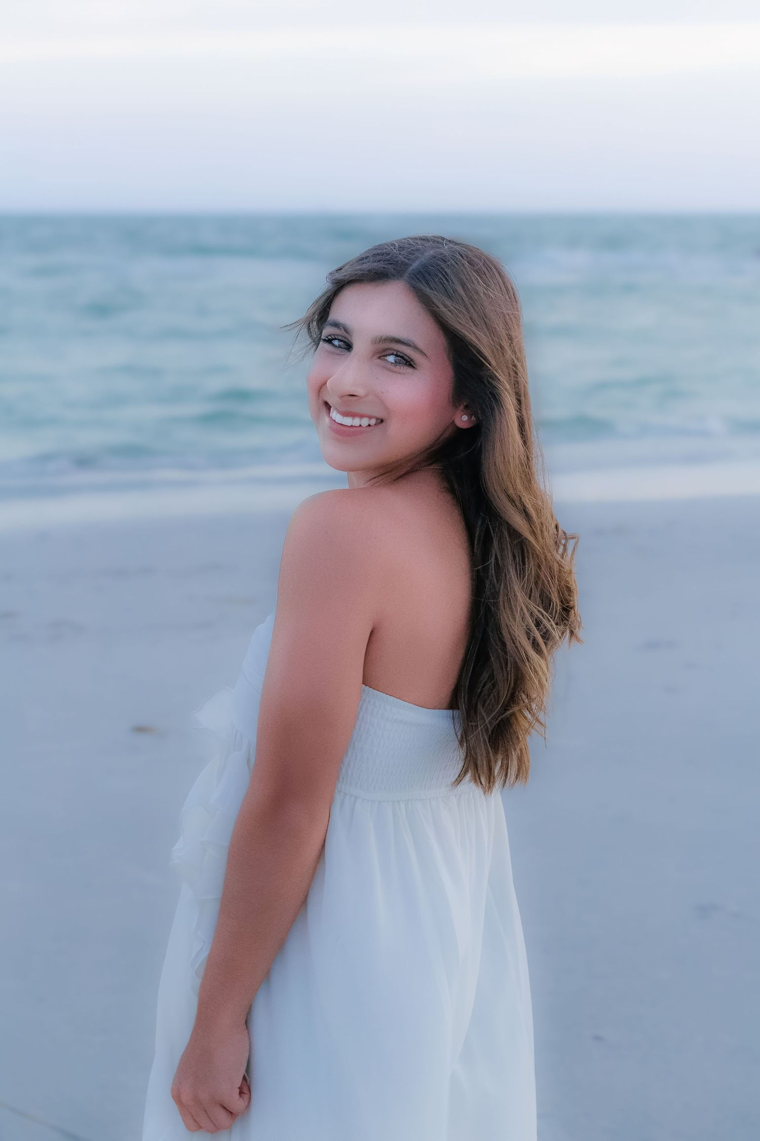 Young woman in white strapless dress smiling over shoulder on sandy beach with turquoise ocean waves behind her.