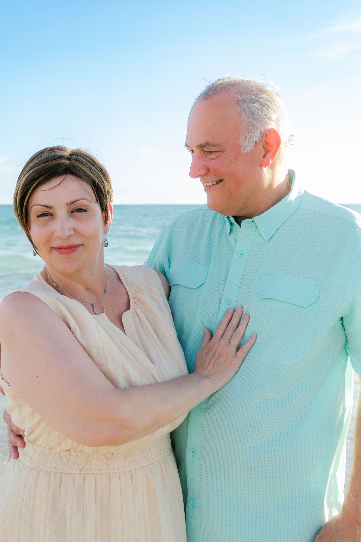 Smiling middle-aged couple embracing on a sunny beach with turquoise ocean waves in the background.