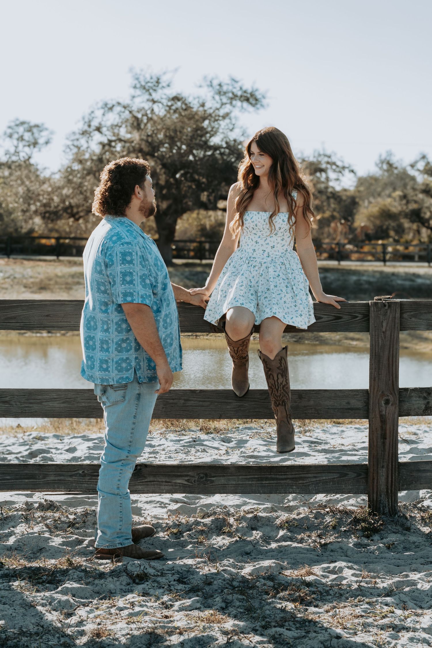 Couple holding hands at a rustic fence, woman in floral dress and cowboy boots, man in blue patterned shirt, outdoor setting.