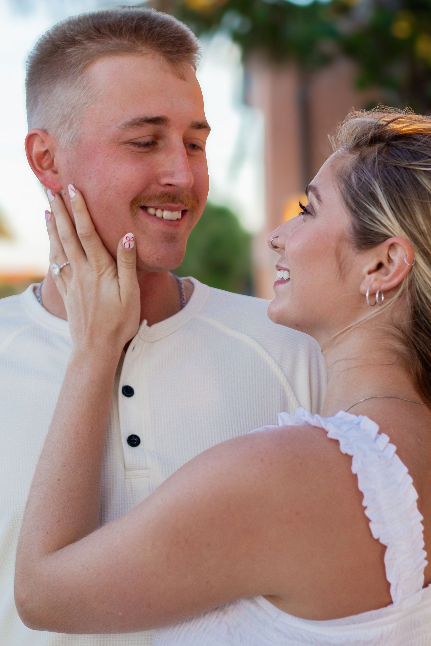A smiling couple in white outfits share a tender moment, with the woman gently touching the man's face.