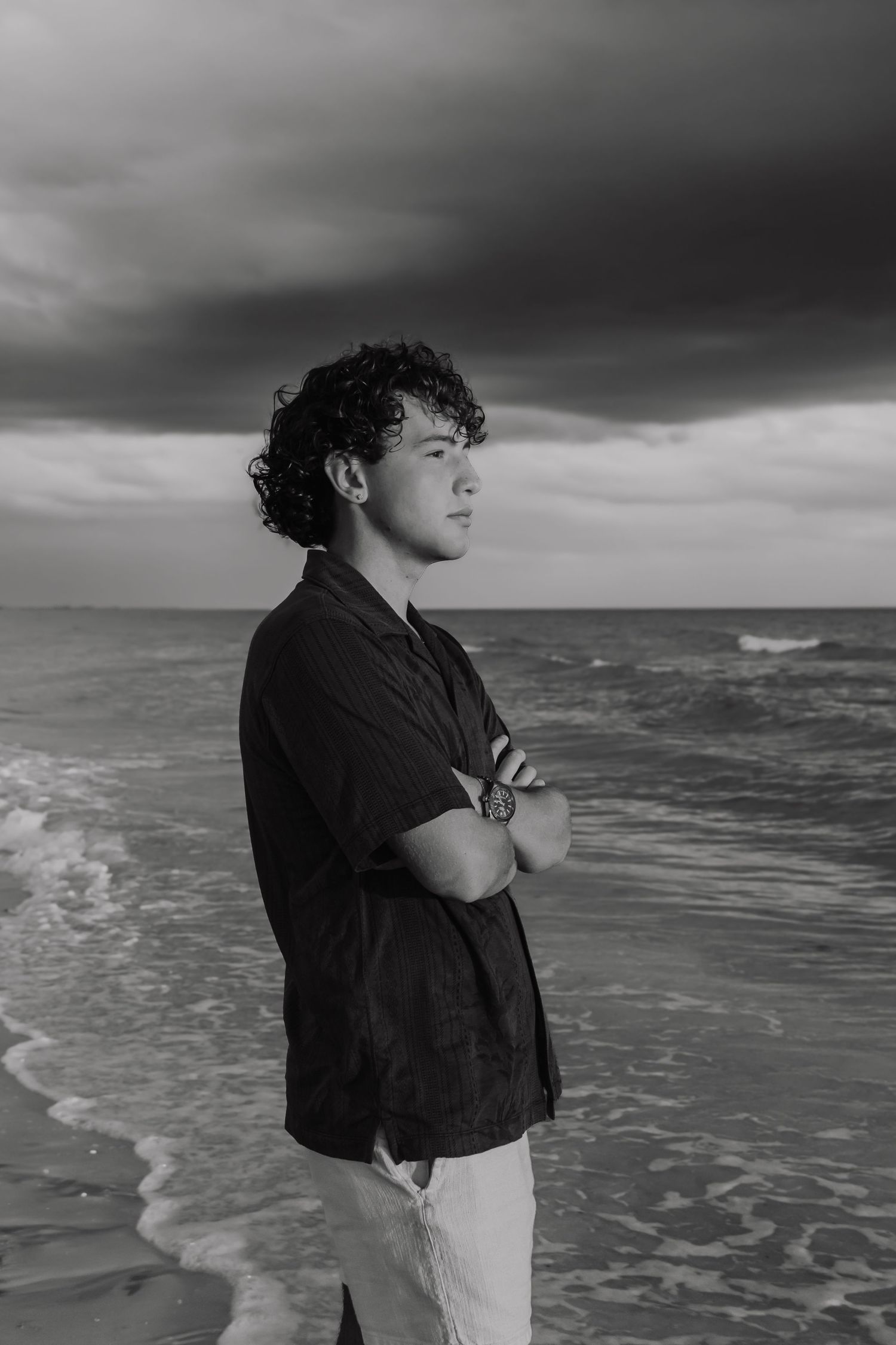 Black and white photo of a young man with curly hair standing on a beach with arms crossed under dramatic cloudy skies.