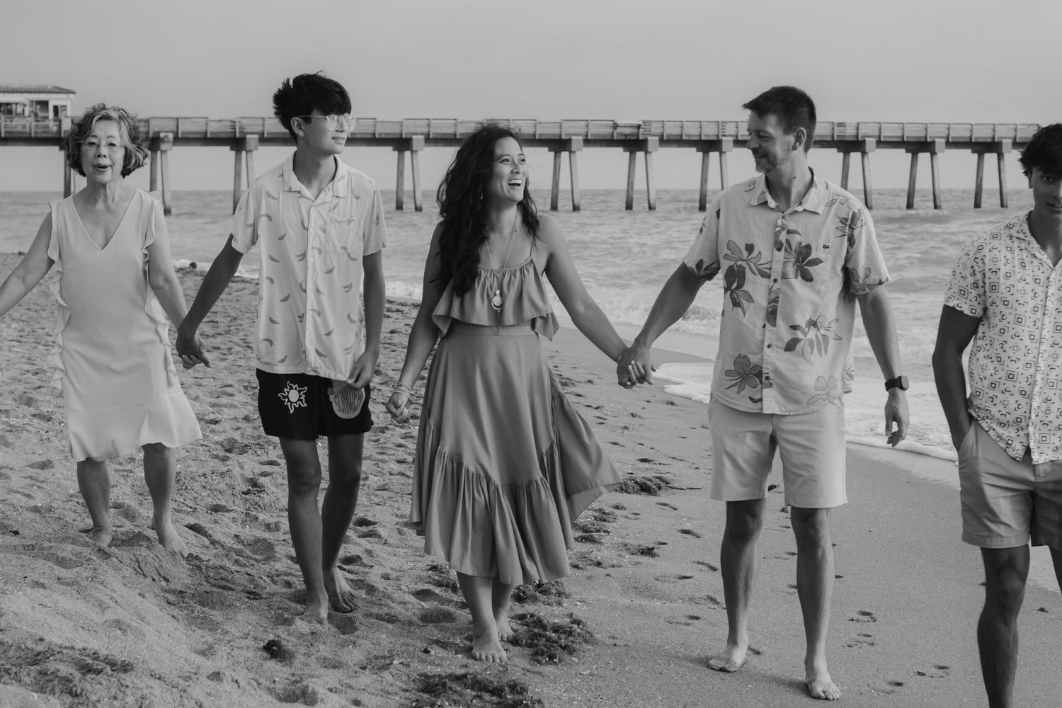 Black and white photo of a family holding hands and walking on a beach near a pier.