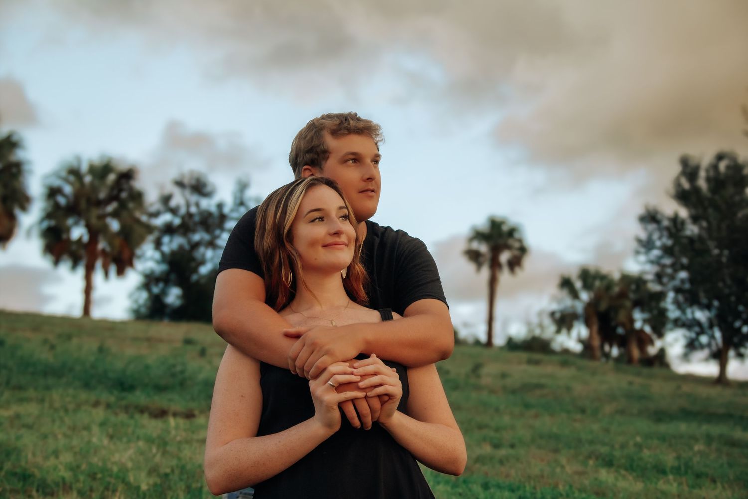 Young couple embracing outdoors on green grass with palm trees and cloudy sky in background.