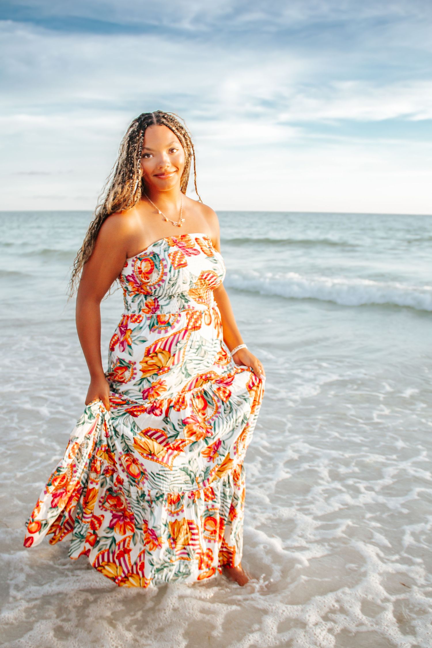 Woman in floral maxi dress standing in ocean waves on a sunny beach.