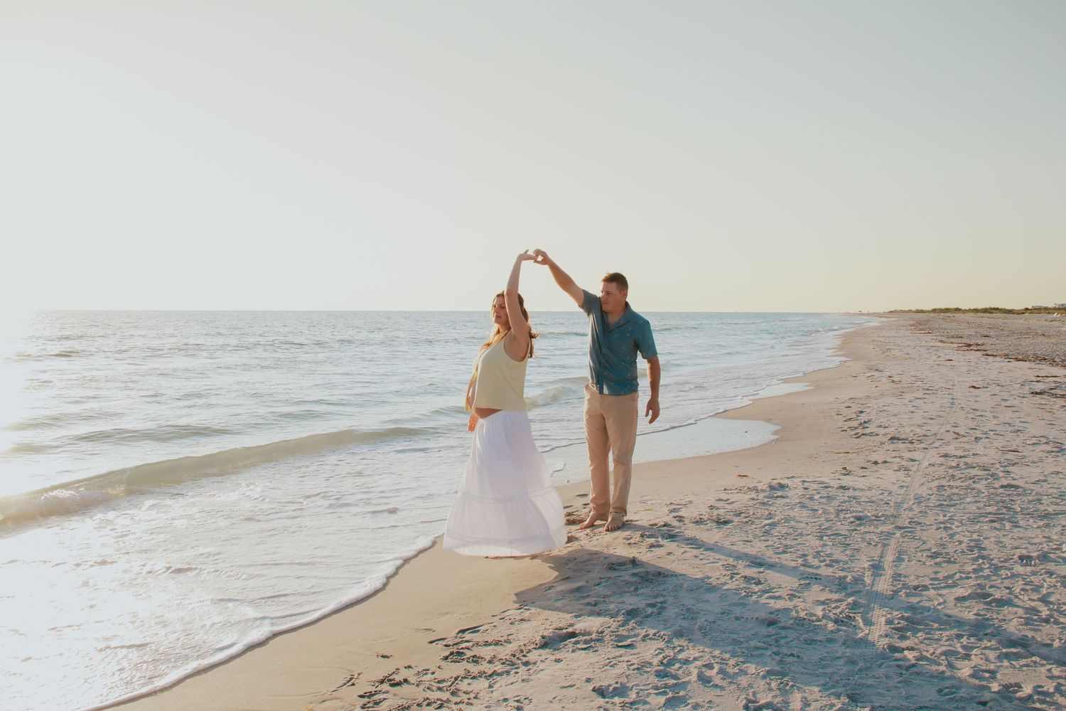 Couple dancing on a sandy beach at sunset, woman in white skirt twirling near ocean waves.