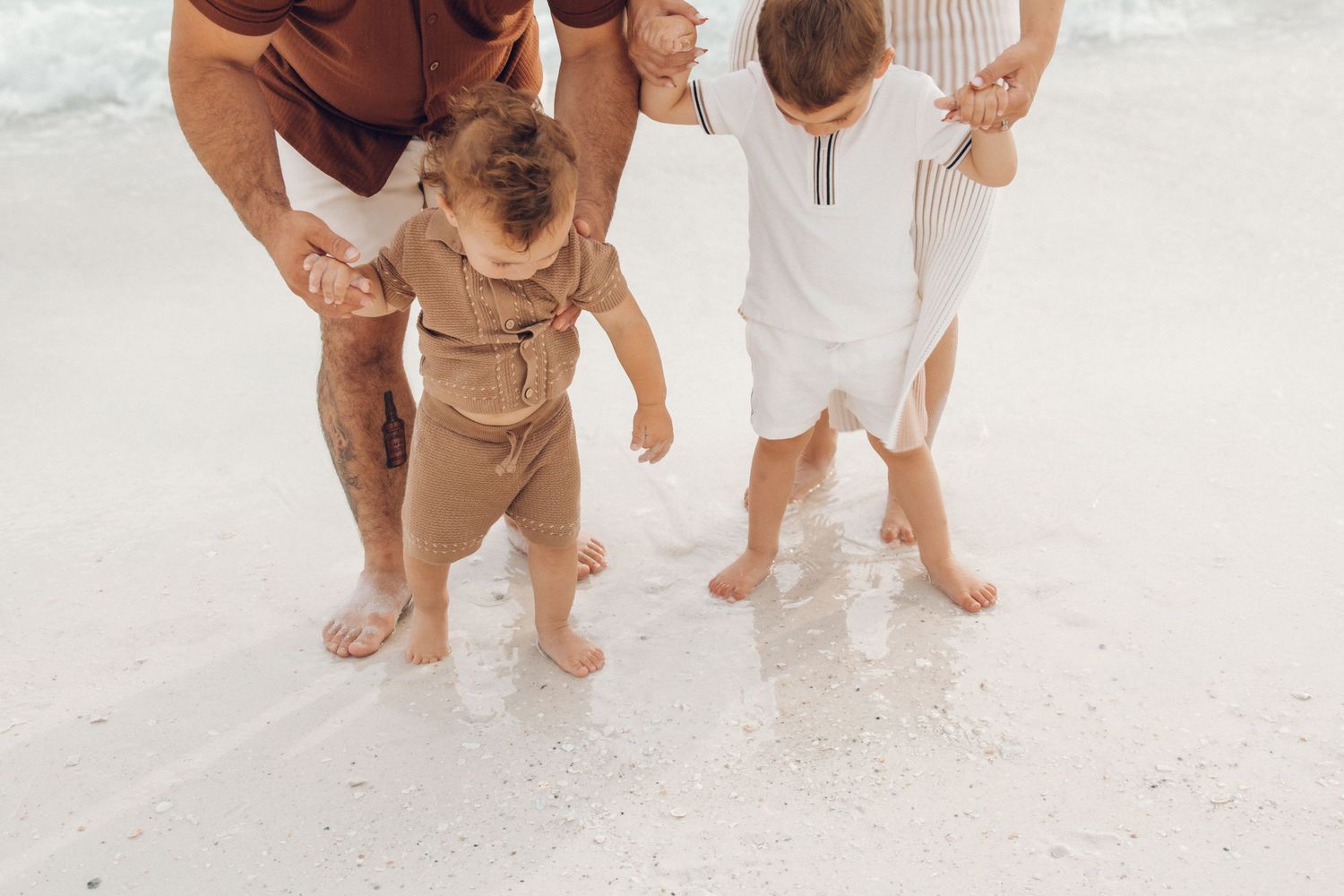 Family with young children standing barefoot in shallow ocean waves on a white sand beach.