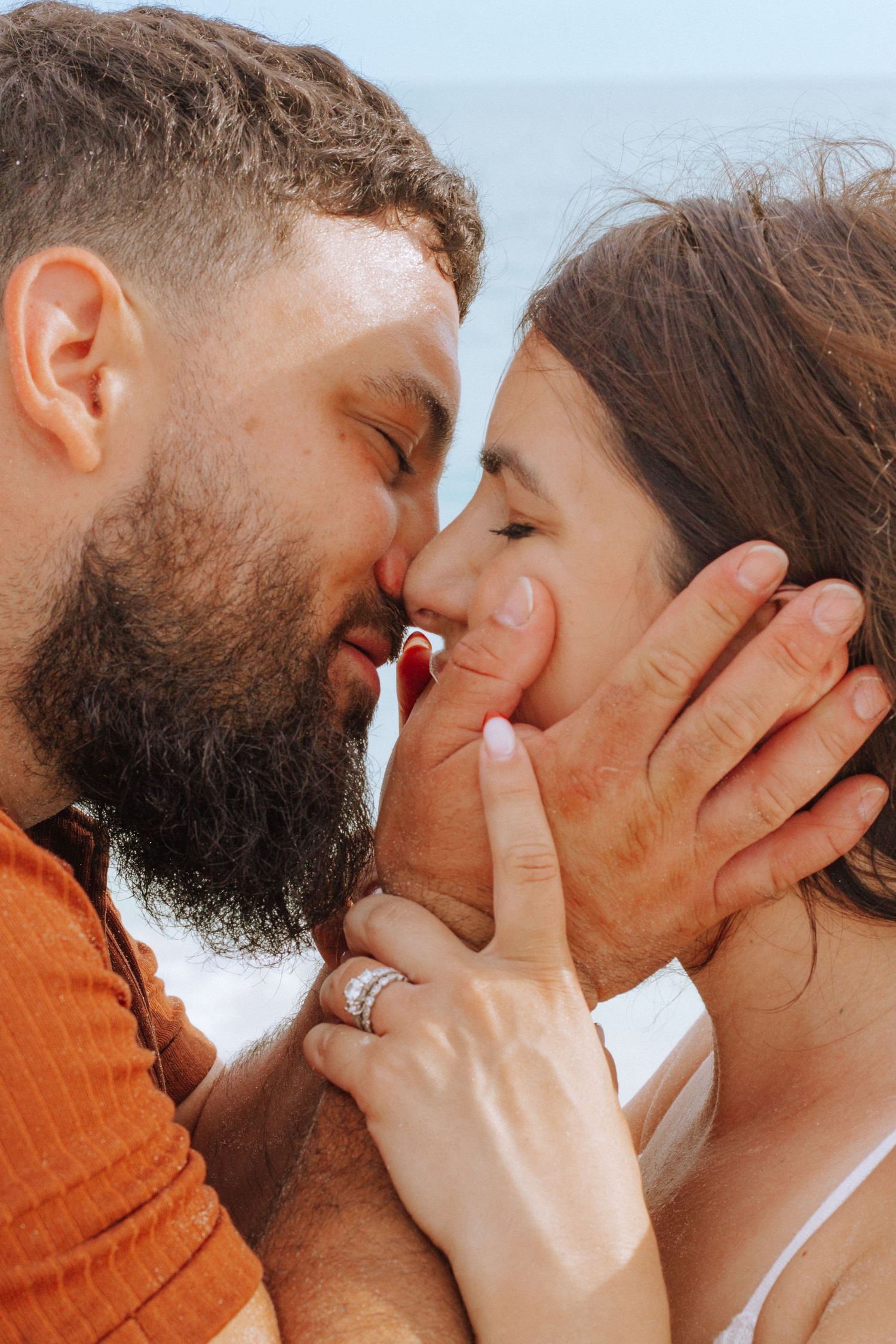 Romantic couple sharing a tender moment outdoors, woman wearing silver rings, man with dark beard, blue sky background.