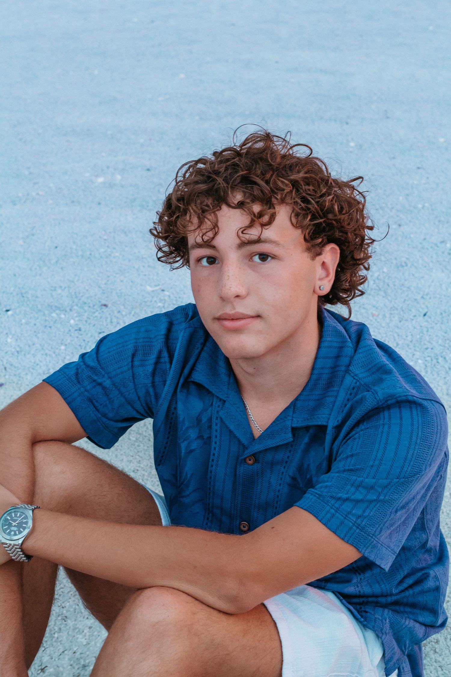 A teenage boy with curly brown hair sits outdoors wearing a blue shirt and watch, posing confidently for a portrait.