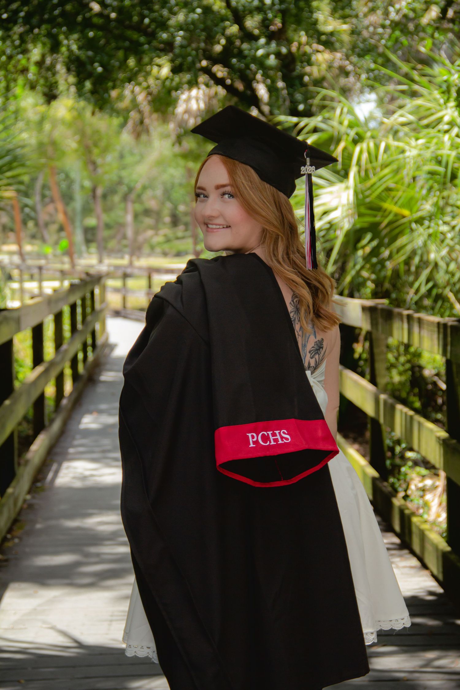 Smiling female graduate in black cap and gown with red PCHS sash poses on wooden bridge surrounded by lush greenery.