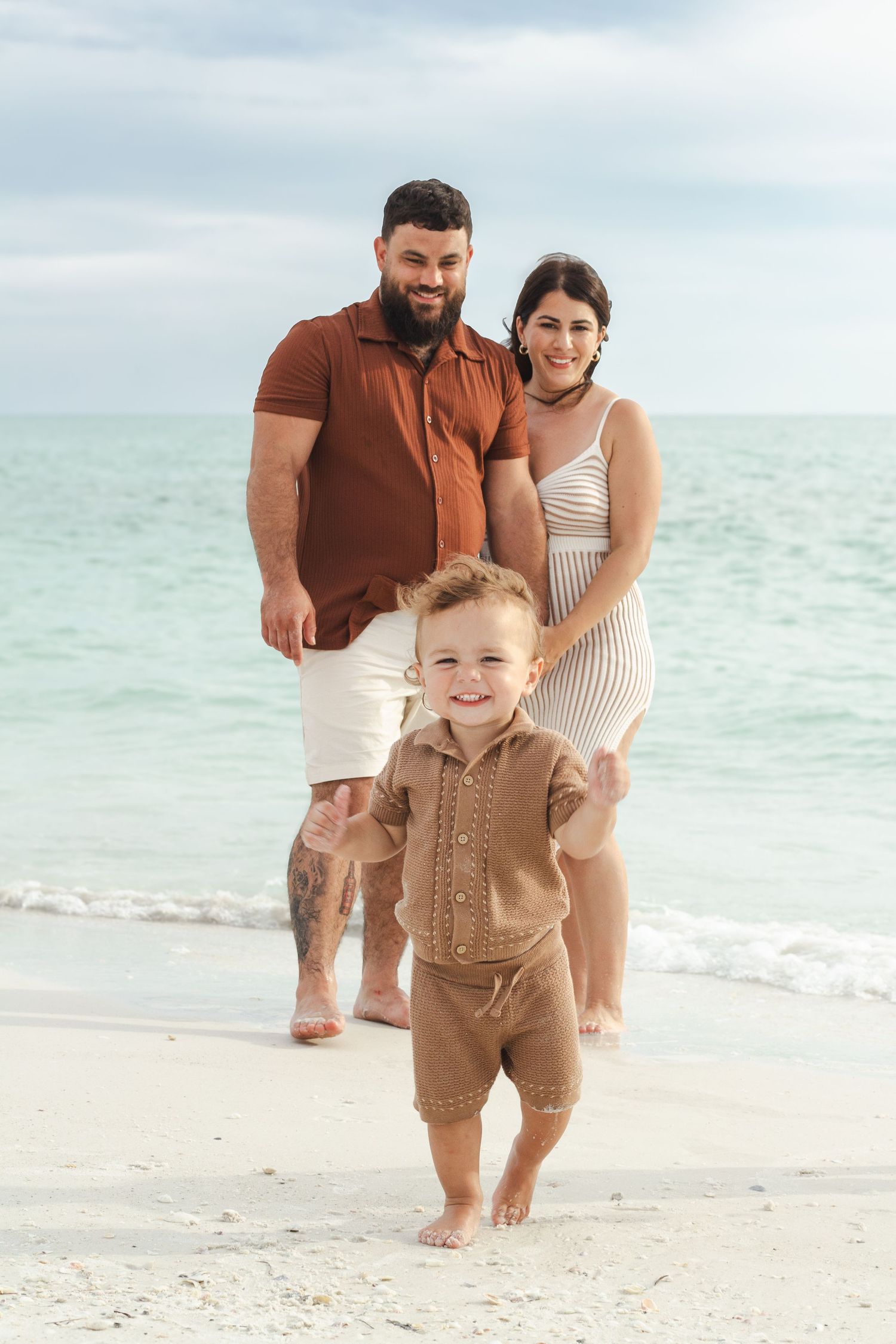 A young family poses on a sandy beach, with parents smiling behind their toddler taking steps toward the camera.