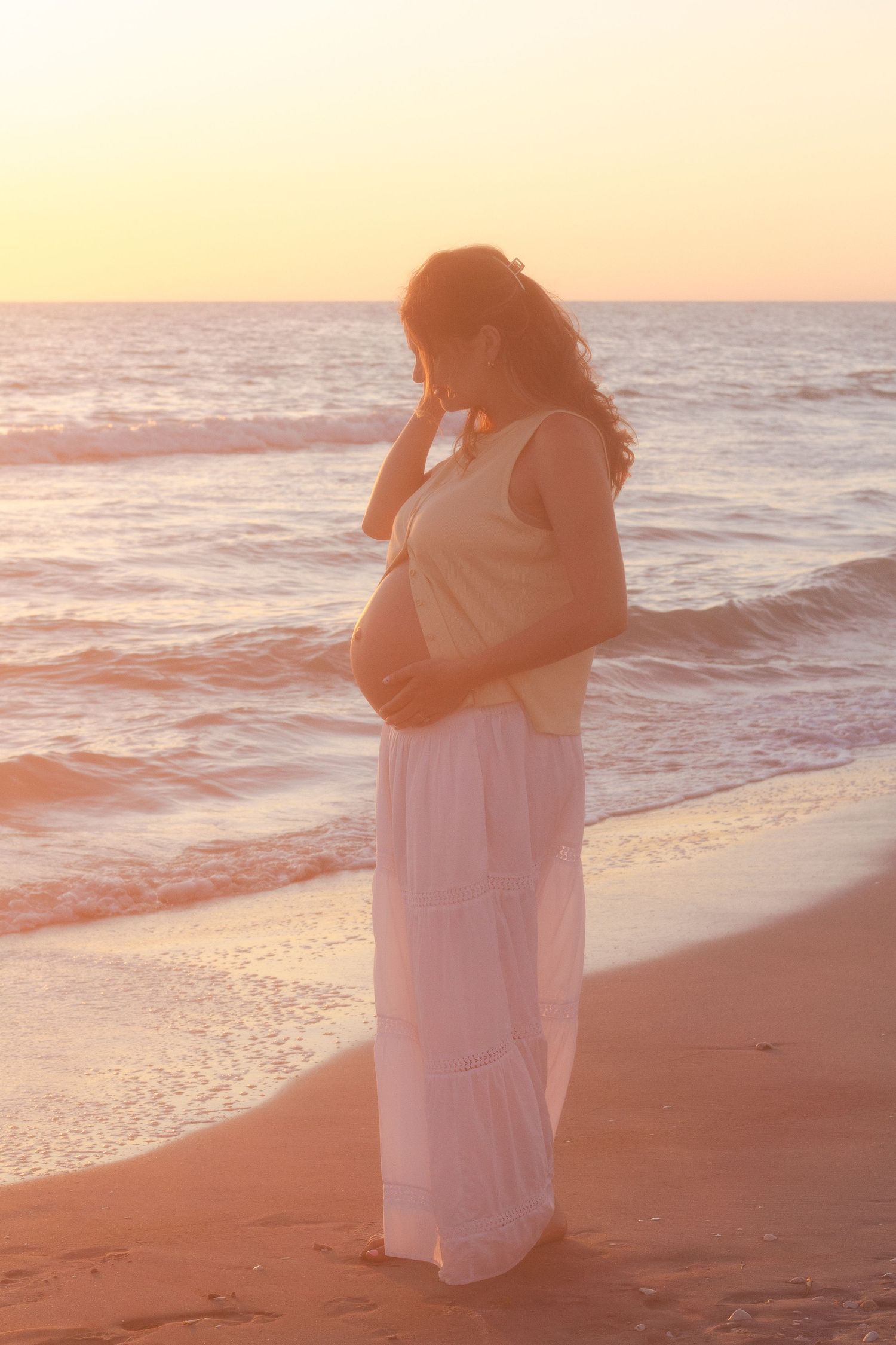 Pregnant woman in white skirt cradling baby bump on sandy beach at golden sunset.
