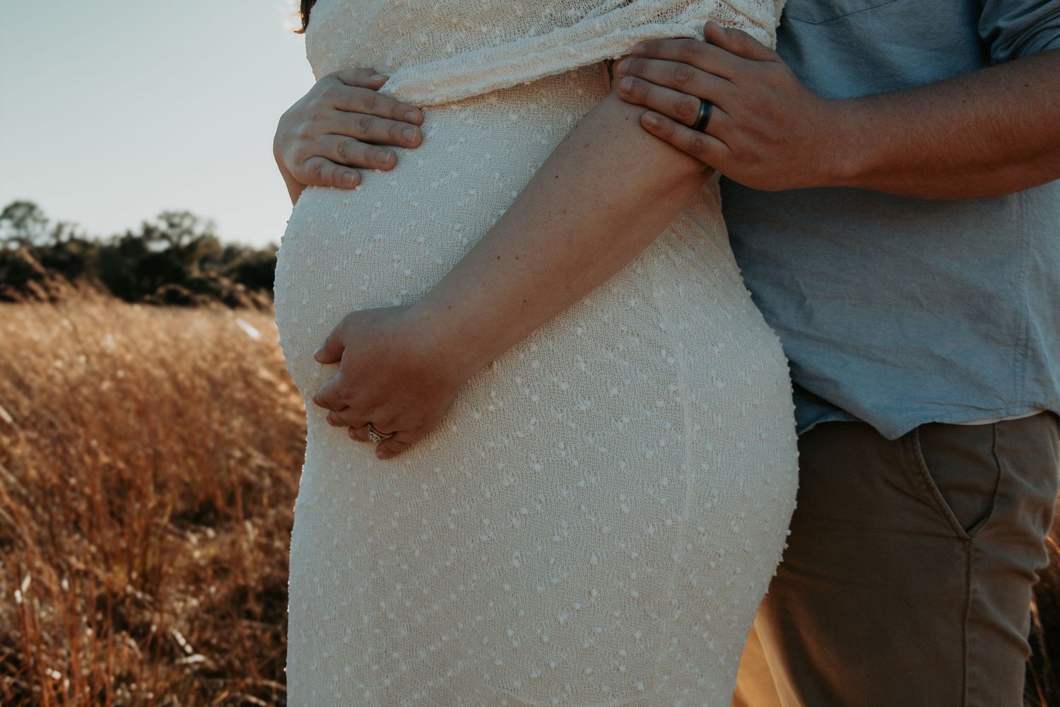 Pregnant couple embracing outdoors in golden field, woman in white lace dress cradling baby bump.