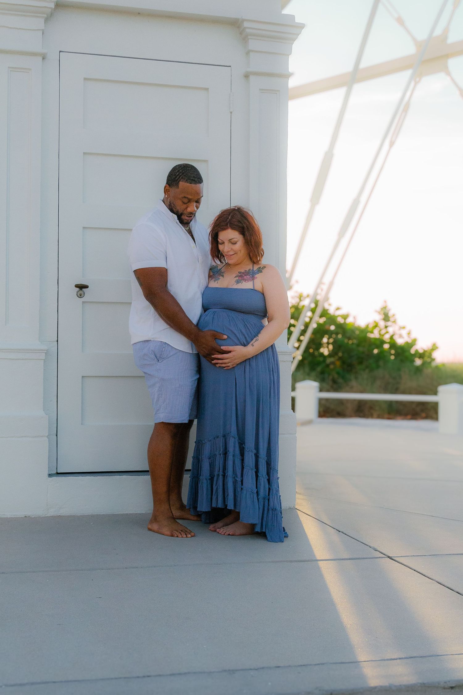 A couple shares a tender maternity moment outdoors, the pregnant woman in a blue dress cradled by her partner.