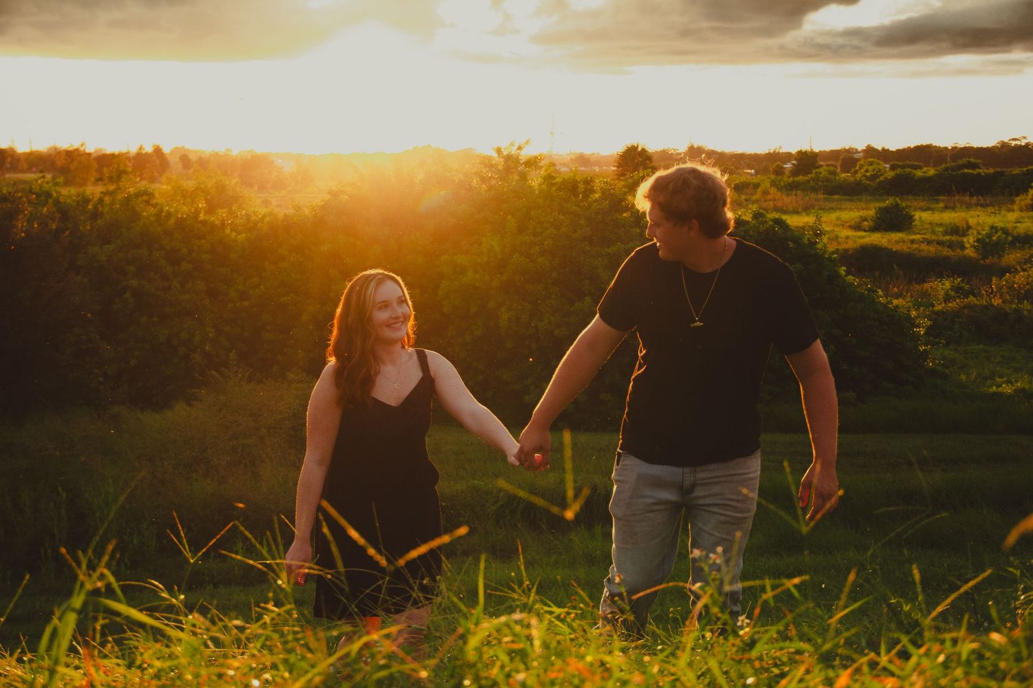 Couple holding hands in a golden sunset field, woman in black dress smiling at man in black shirt.