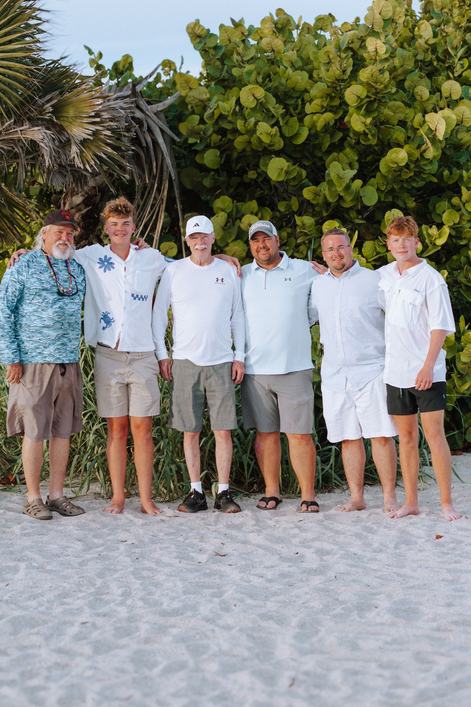 Six men posing together on a white sand beach with tropical greenery backdrop on a sunny day.