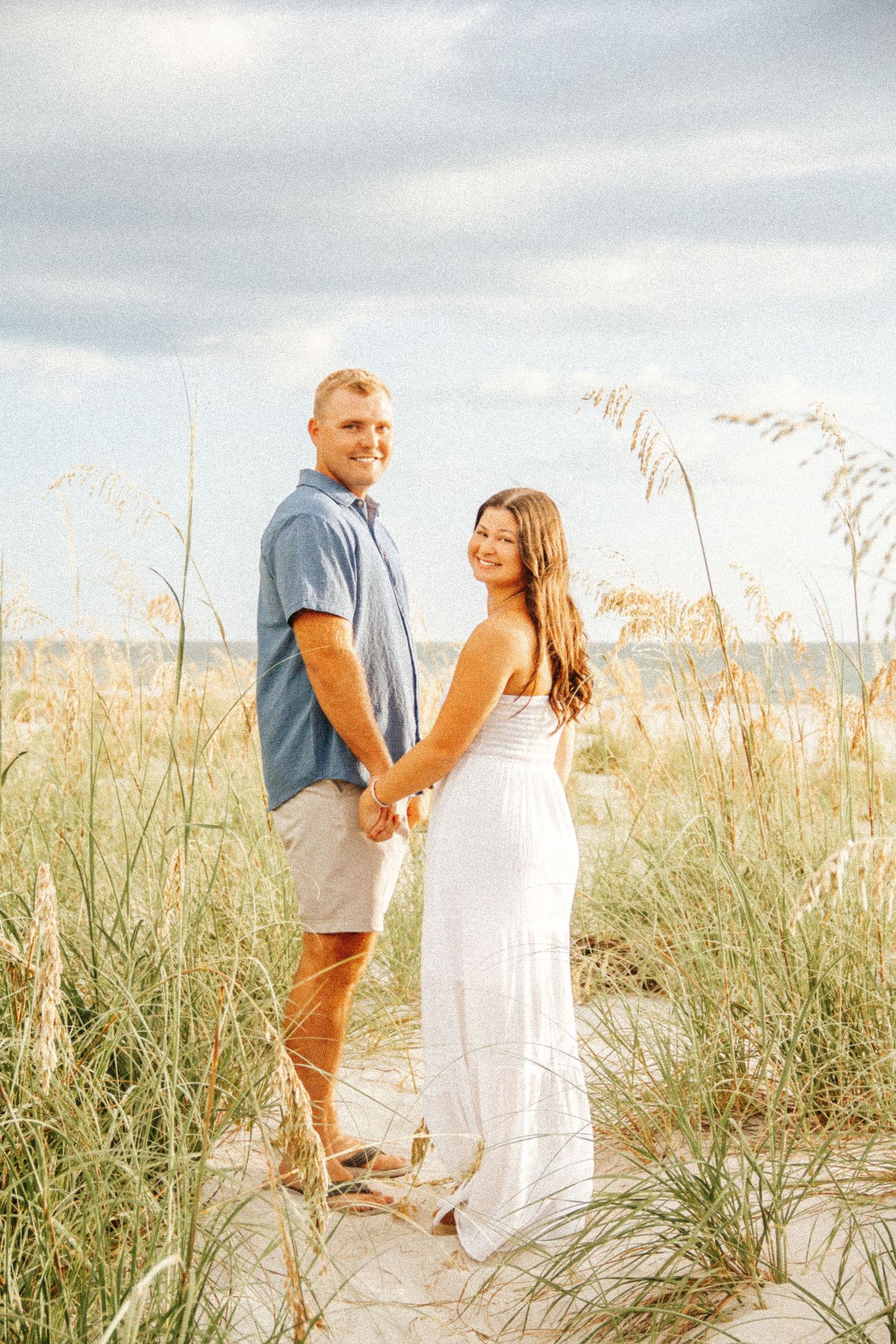 Couple holding hands on sandy beach dunes with sea oats, woman in white dress, man in blue shirt, ocean backdrop.
