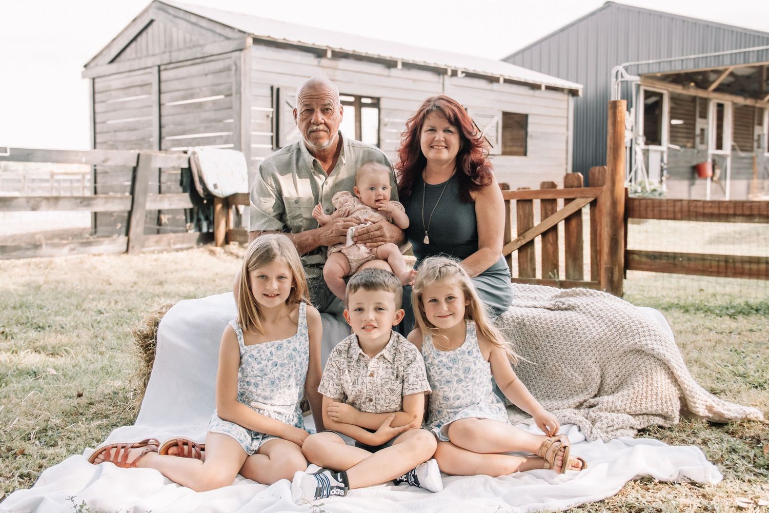 Multi-generational family portrait with grandparents and four young children on a blanket at a rustic farm setting.
