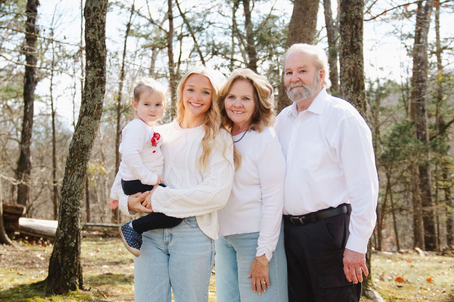 Family of four posing outdoors in white outfits among trees, including grandparents and young child.