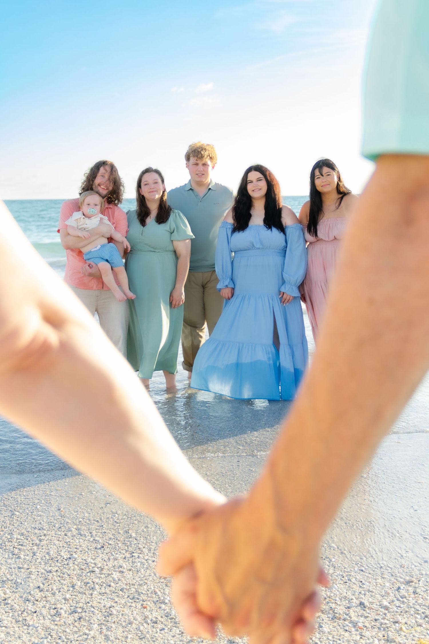 Family group of six adults and a baby holding hands on a sandy beach with clear blue skies.