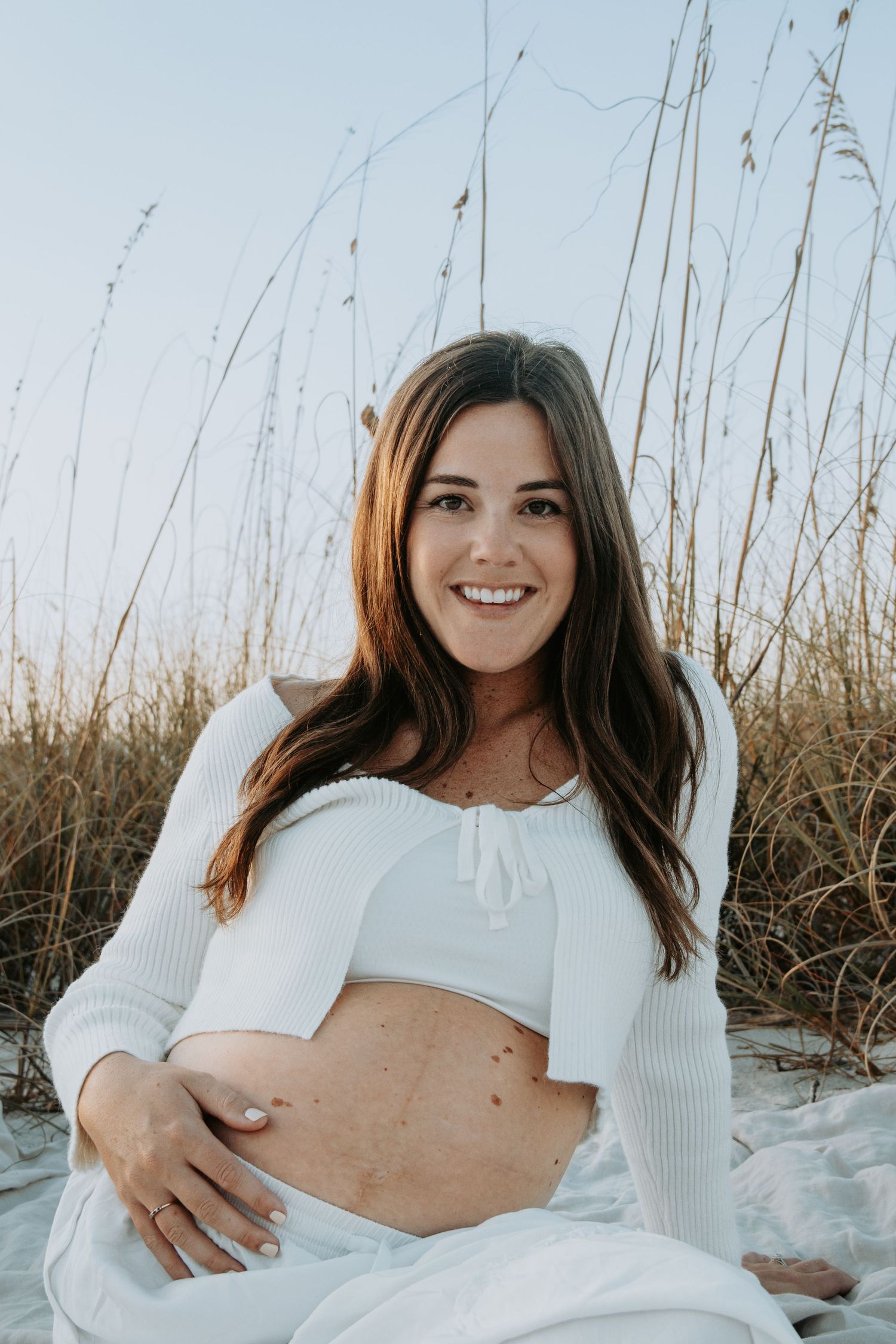 Smiling pregnant woman in white crop top sitting on beach blanket among tall sea grass during golden hour.