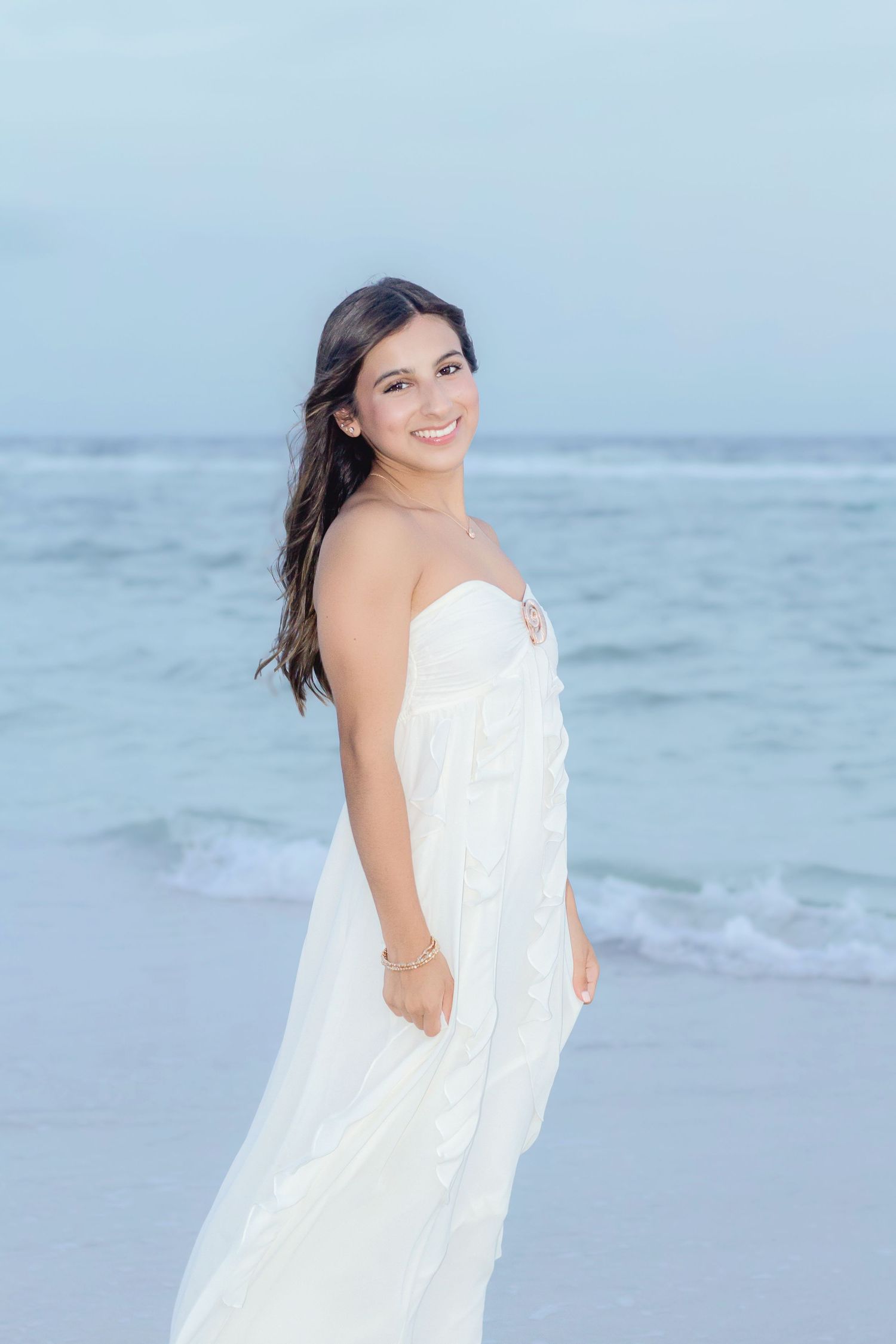 Young woman in white strapless dress smiling on a beach with ocean waves in the background.