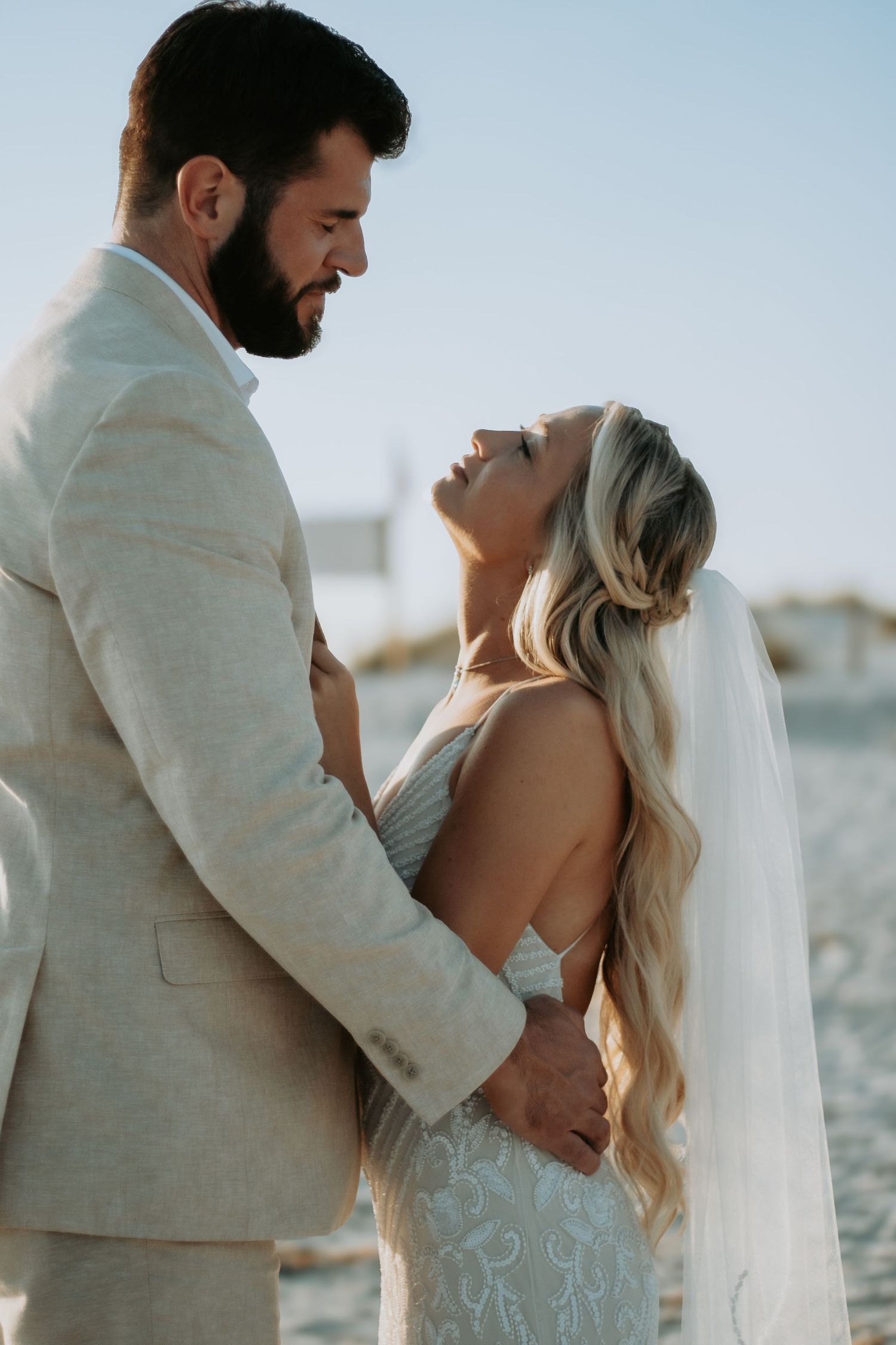 Bride and groom share a romantic beach moment, she in a lace gown with veil, he in a tan suit.
