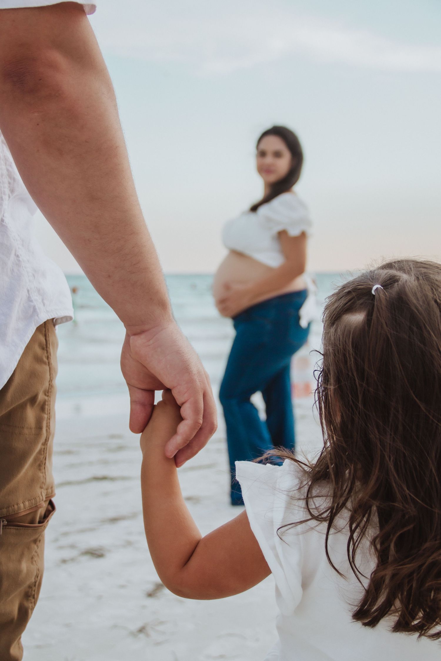 A young girl holds her father's hand on a beach while her pregnant mother stands in the background near the ocean.