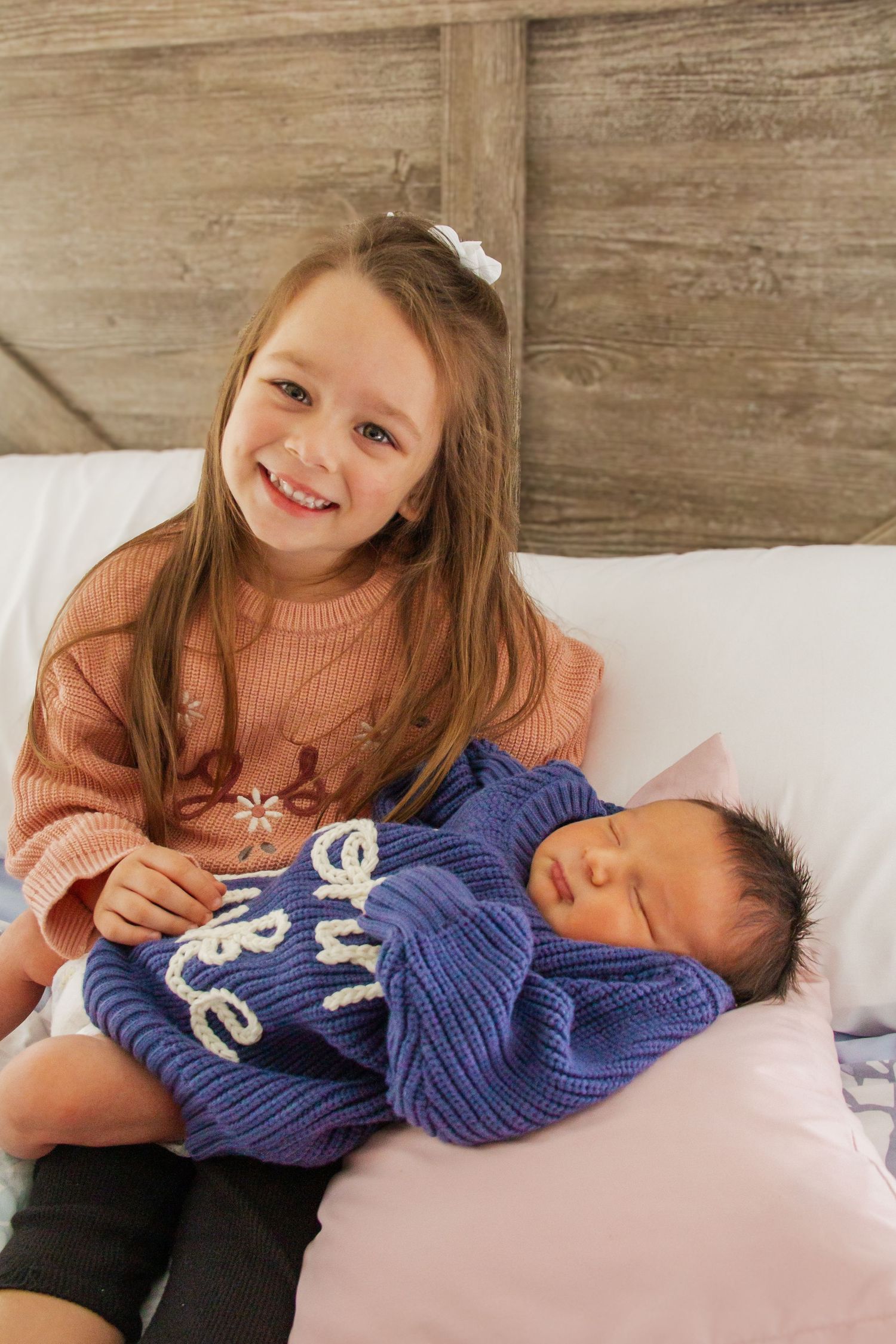 Young girl smiling while holding a sleeping newborn baby wrapped in a blue knit sweater on a bed.