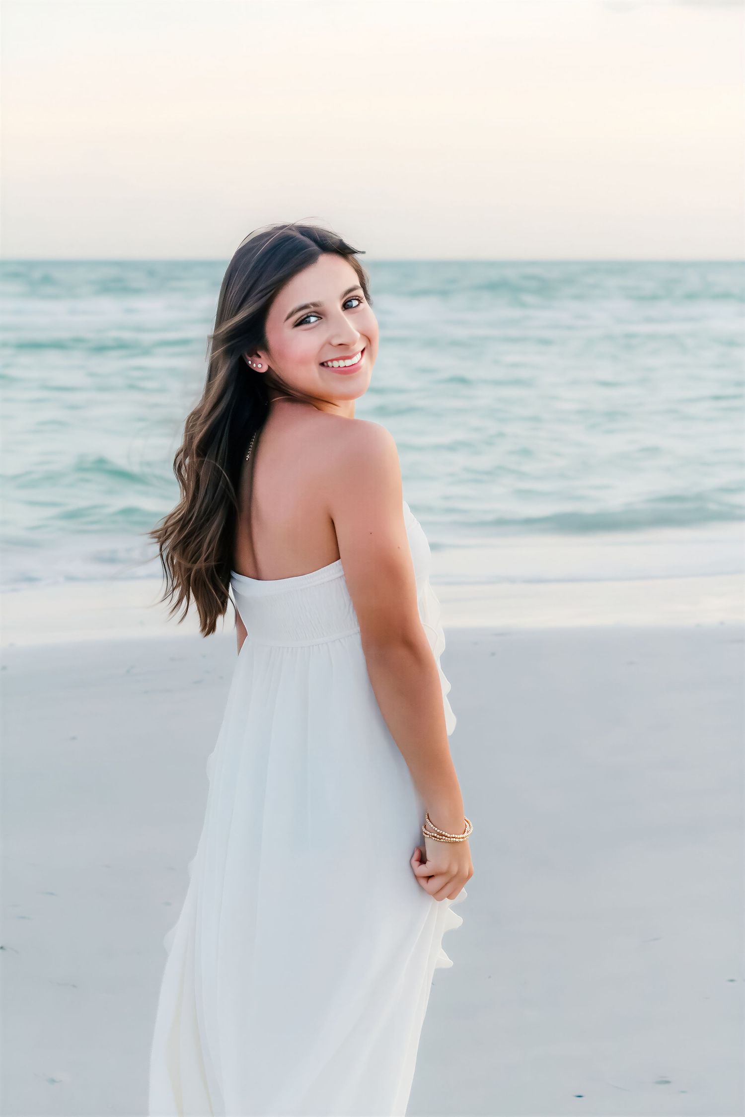 Young woman in white strapless dress smiling on a sandy beach with turquoise ocean waves behind her.