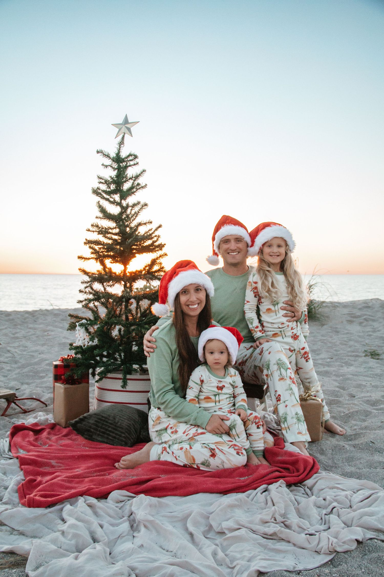 A festive beach photo session with a small decorated Christmas tree and red blanket on the sand at sunset.