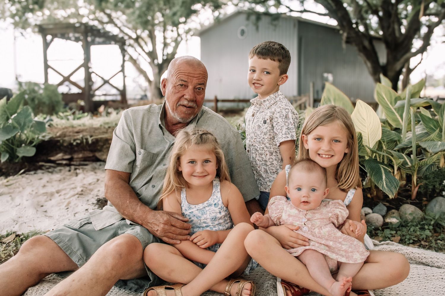 Grandfather smiling with four young grandchildren outdoors near a beach cottage surrounded by tropical plants.