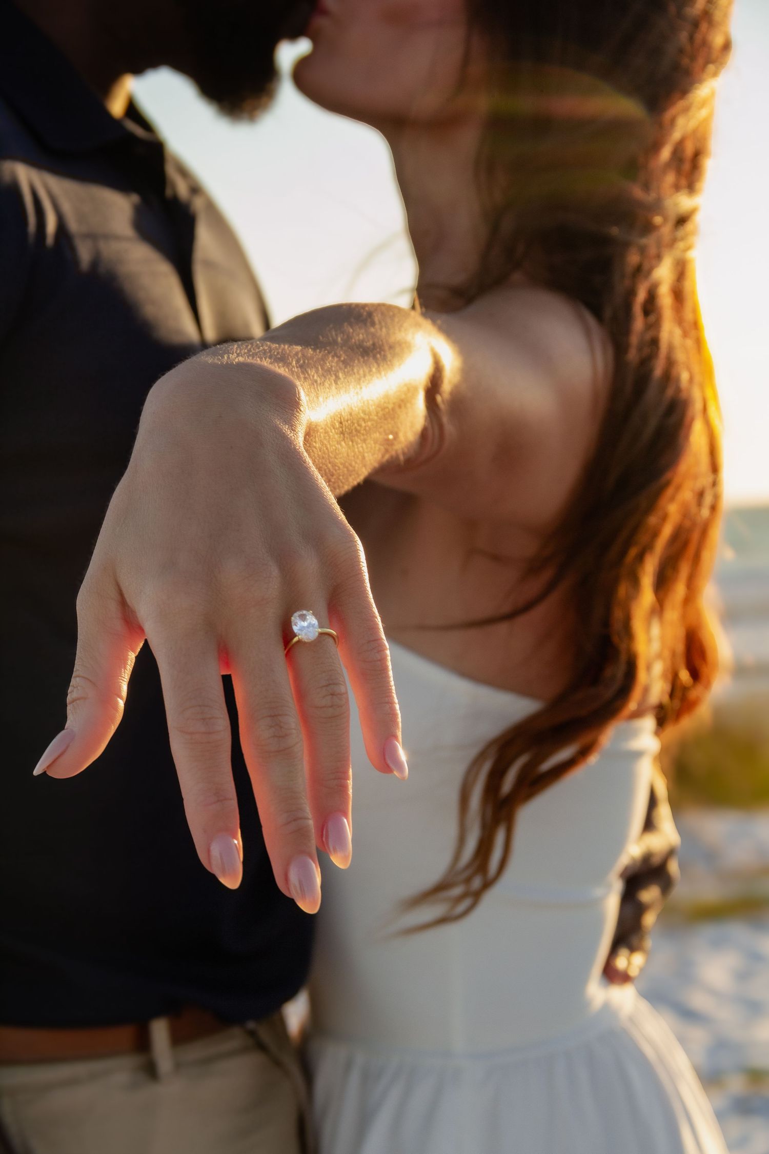 Couple sharing a kiss at sunset while showing off a stunning diamond engagement ring on her outstretched hand.
