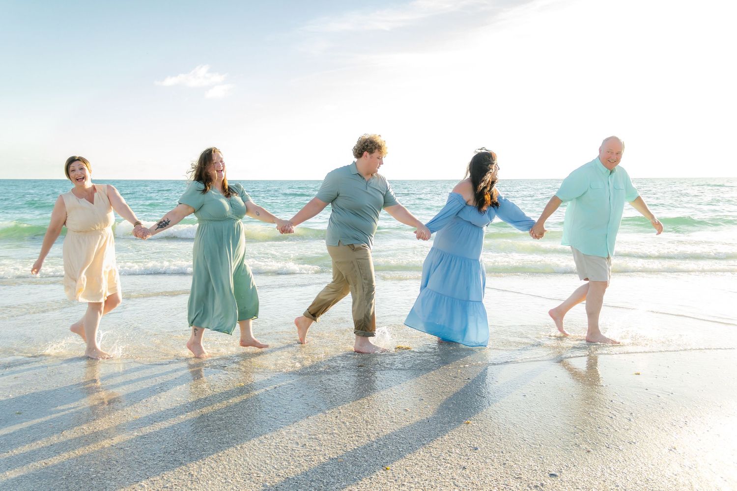 Five people hold hands walking along a sunny beach shore in pastel outfits, waves reflecting golden light behind them.
