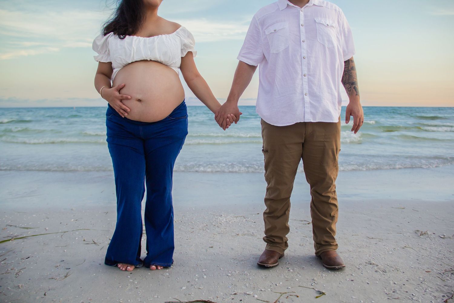 Pregnant couple holding hands on a beach at sunset, woman in white top and blue jeans cradling baby bump.