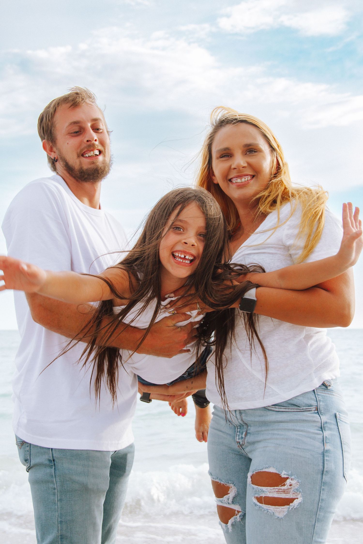 Happy family in white shirts and jeans playing on the beach, parents lifting smiling daughter with ocean waves behind them.
