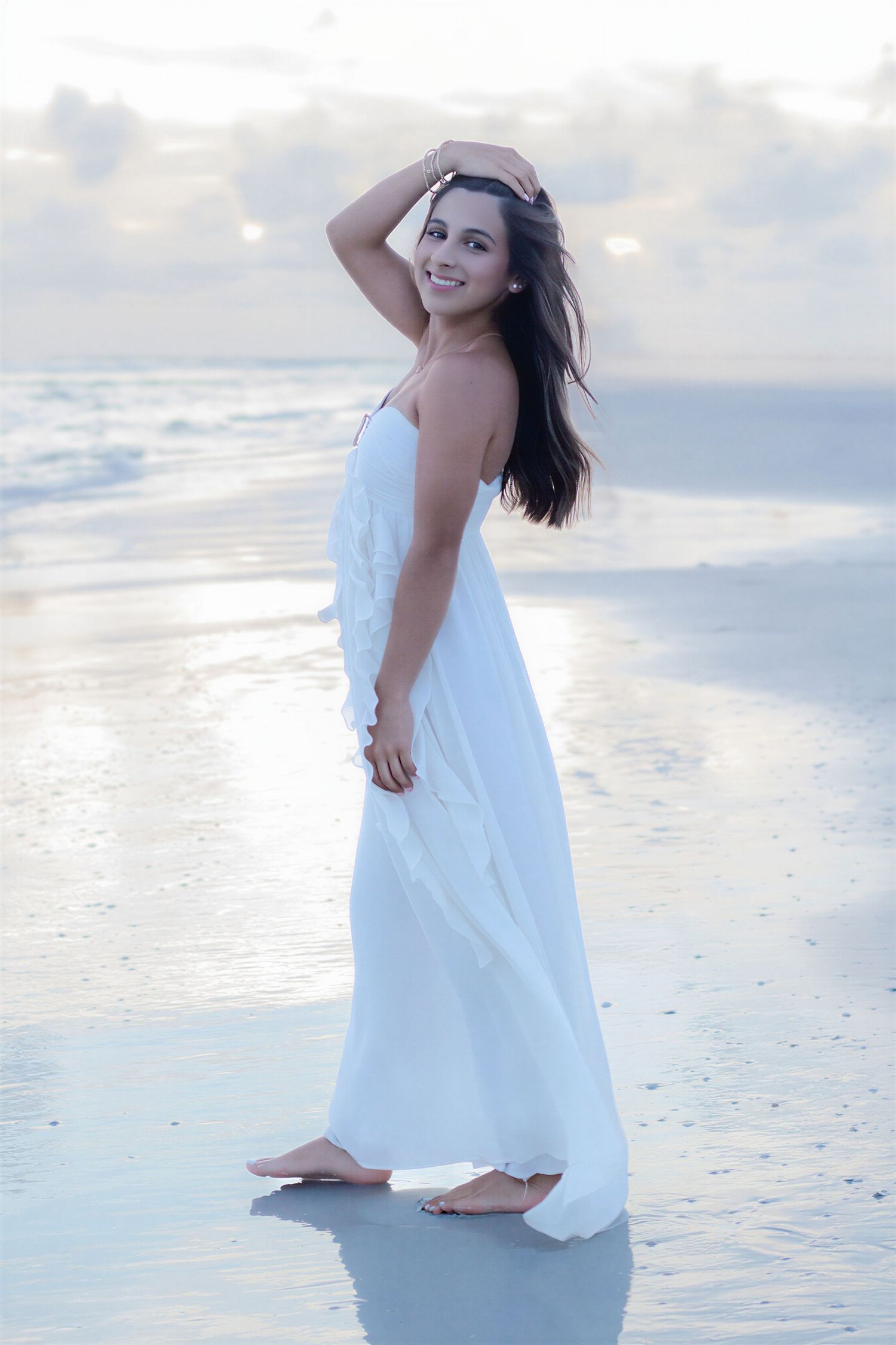Young woman in flowing white dress standing barefoot on a serene beach at sunset, smiling with hand in hair.