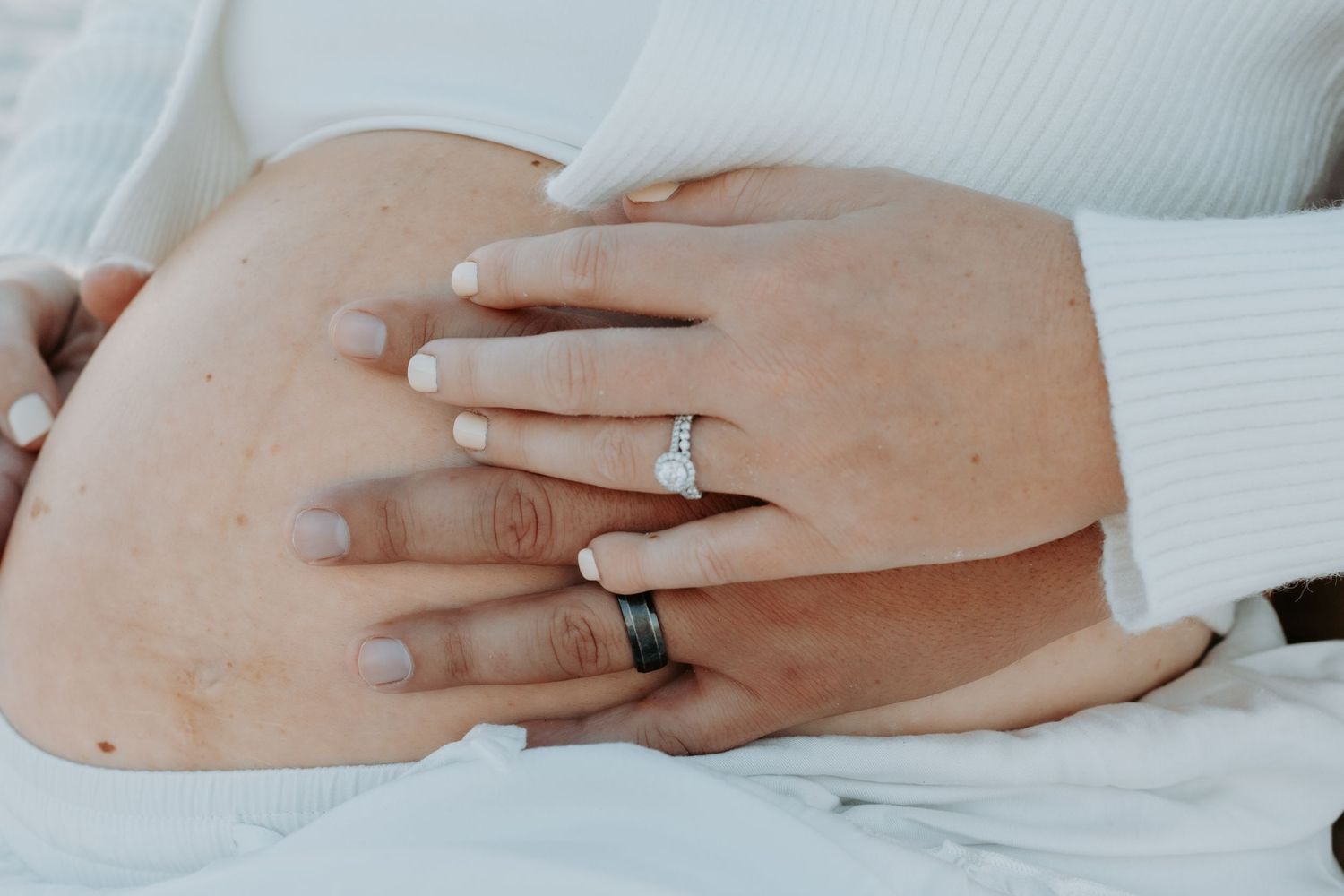 Close-up of couple's hands with diamond engagement ring and black wedding band resting on pregnant belly.
