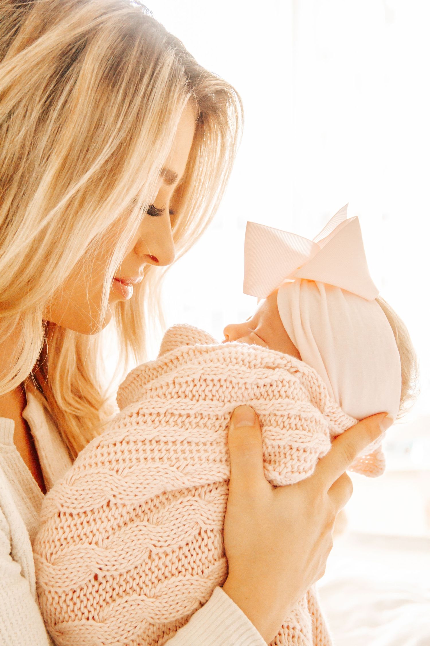 A blonde mother tenderly holds her newborn baby wrapped in a soft pink knit blanket with a bow headband.