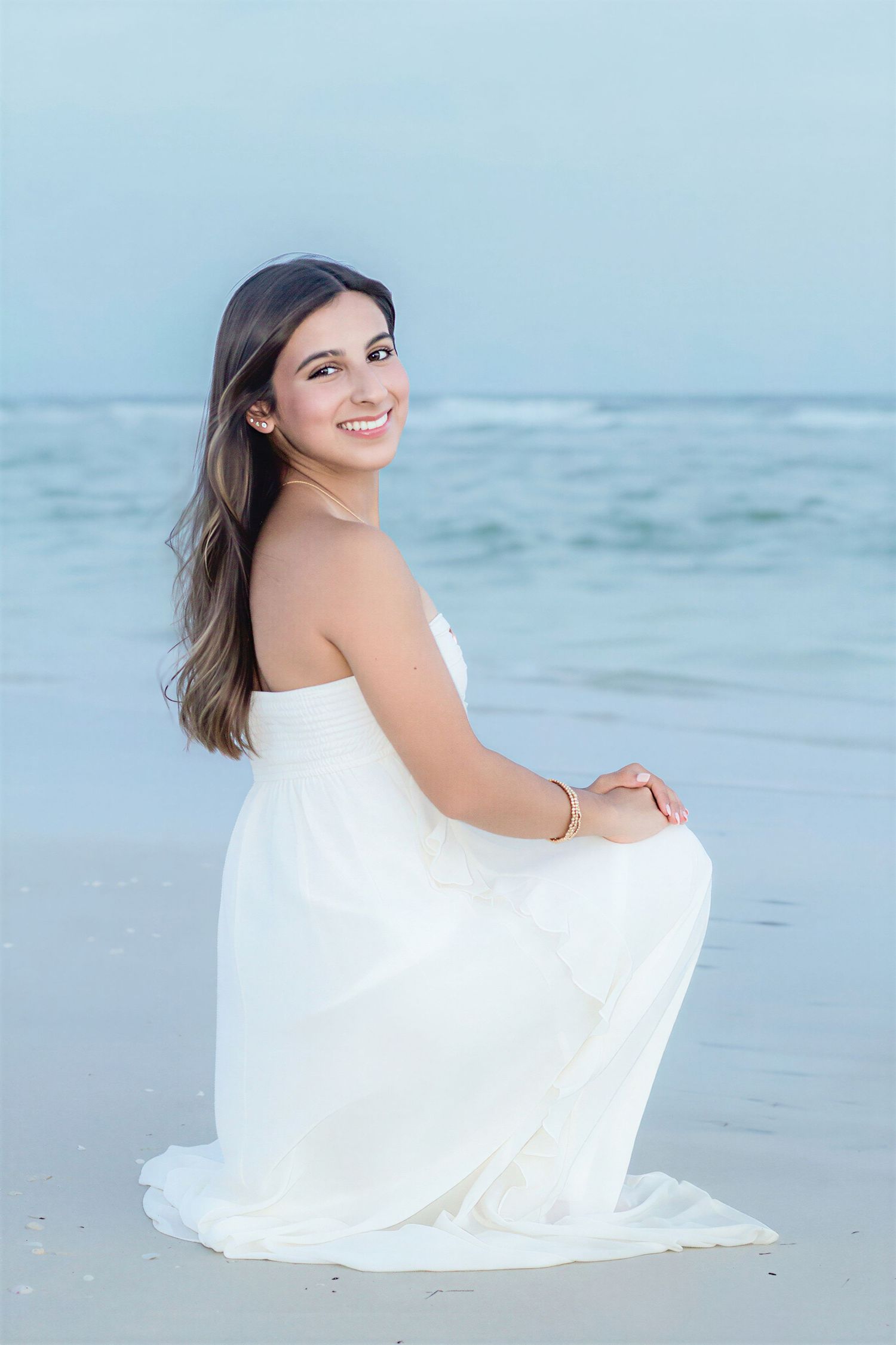 Young woman in white strapless gown kneeling on sandy beach, smiling, with ocean waves in background.