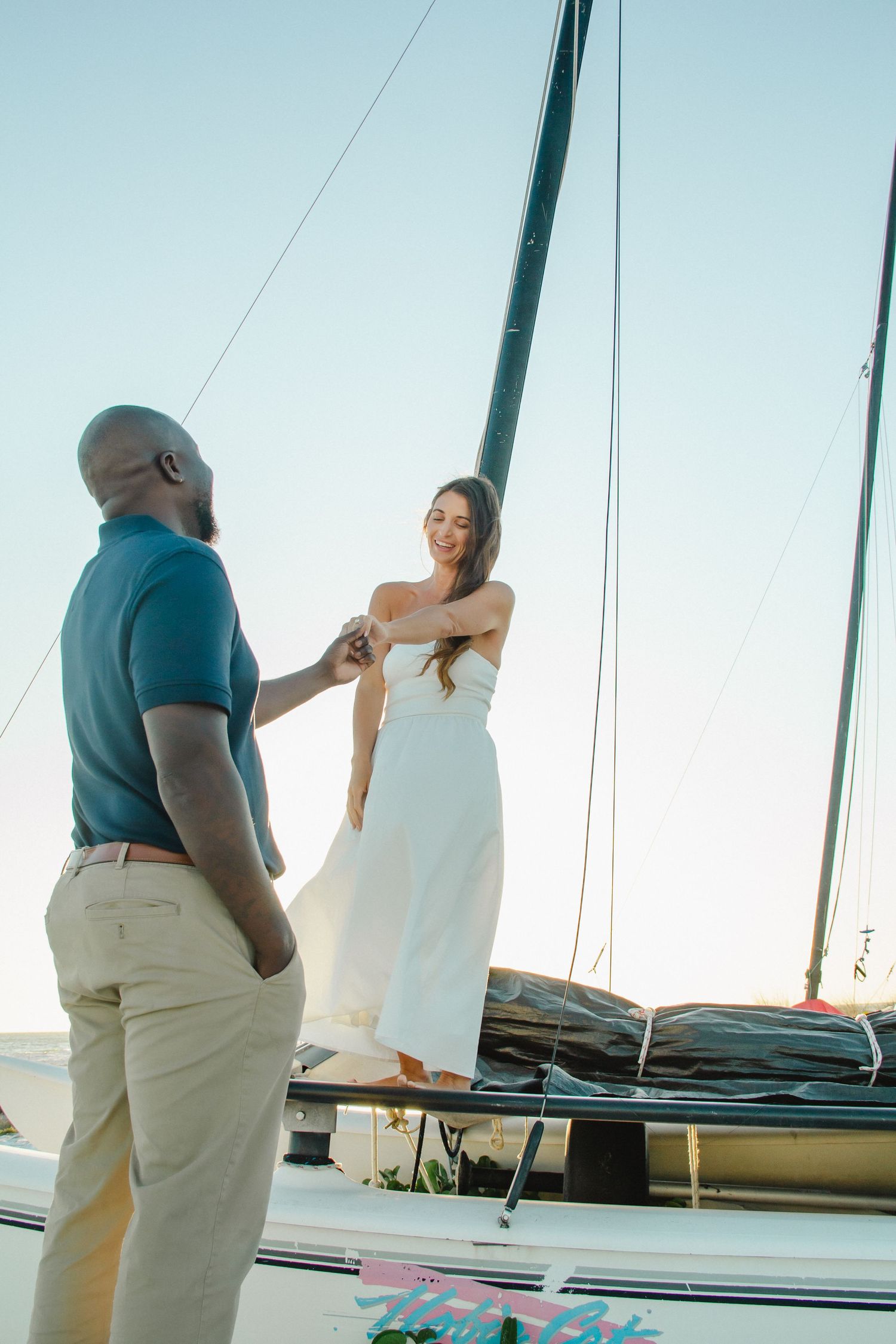 Interracial couple holding hands on a sailboat at sunset, woman in white dress smiling.