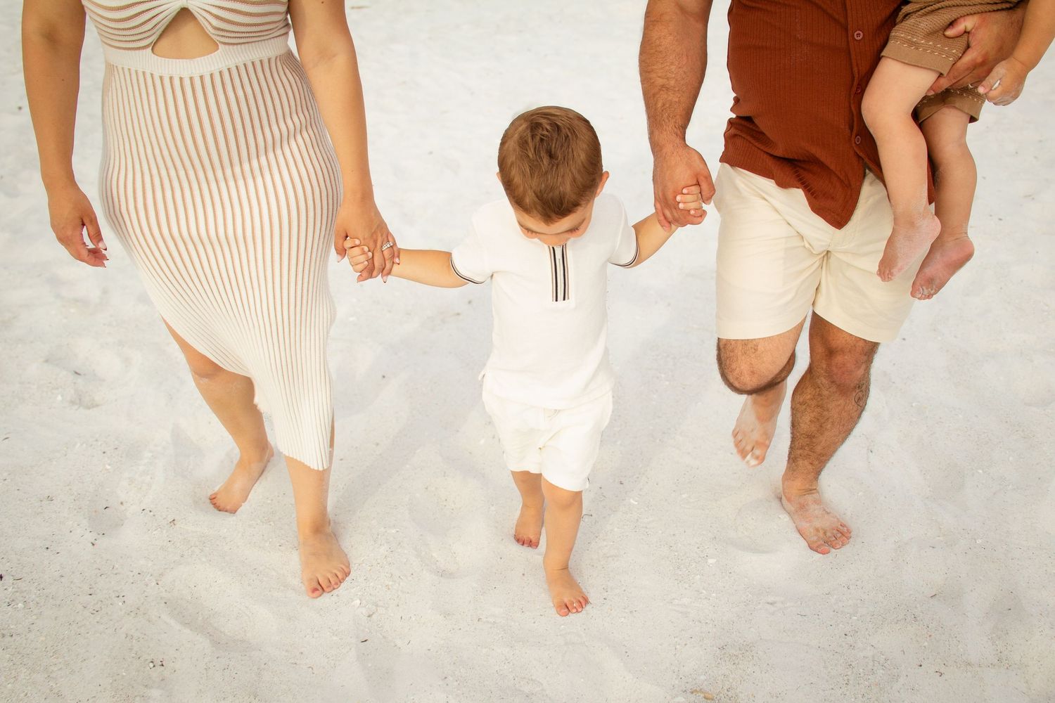 Family of four walking barefoot on white sand beach, parents holding toddler's hands while carrying baby.