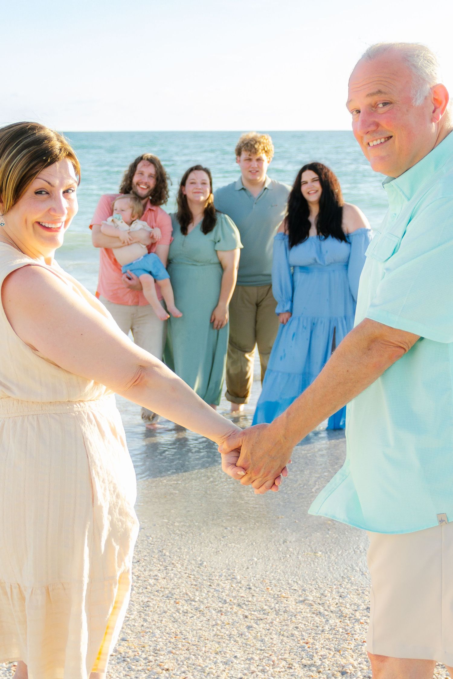 Family group photo on a sunny beach, with couple holding hands in foreground and five relatives standing in ocean waves behind them.