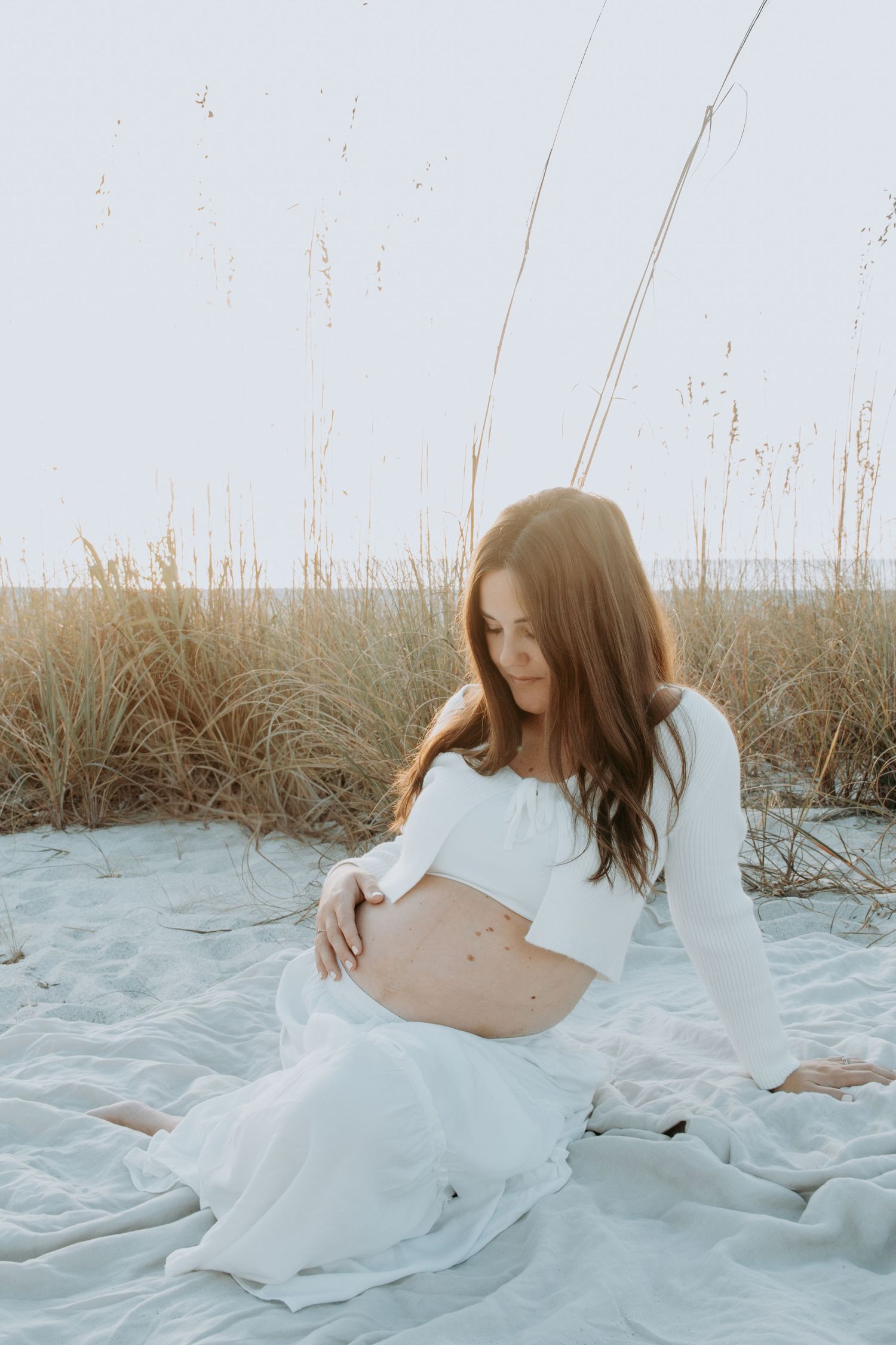 Pregnant woman in white crop top and skirt sitting on beach blanket among sea oats, cradling her baby bump.