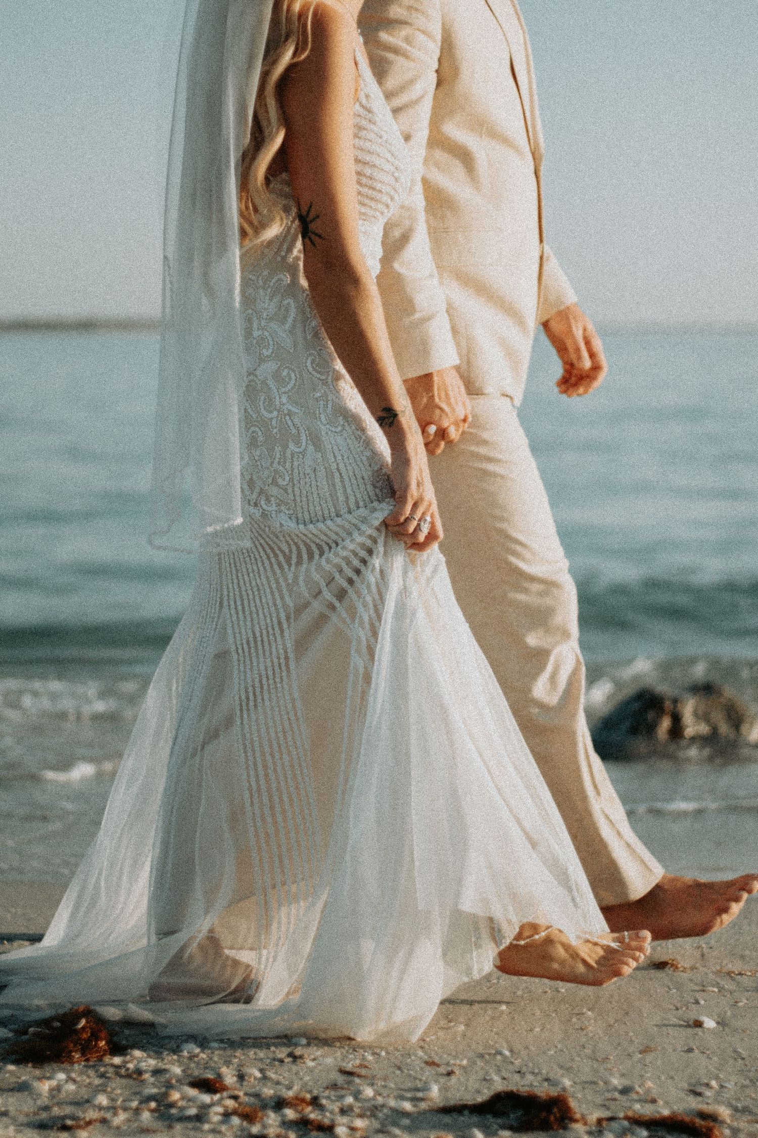 Bride and groom walking barefoot on beach, wearing white gown and beige suit, holding hands by ocean waves.
